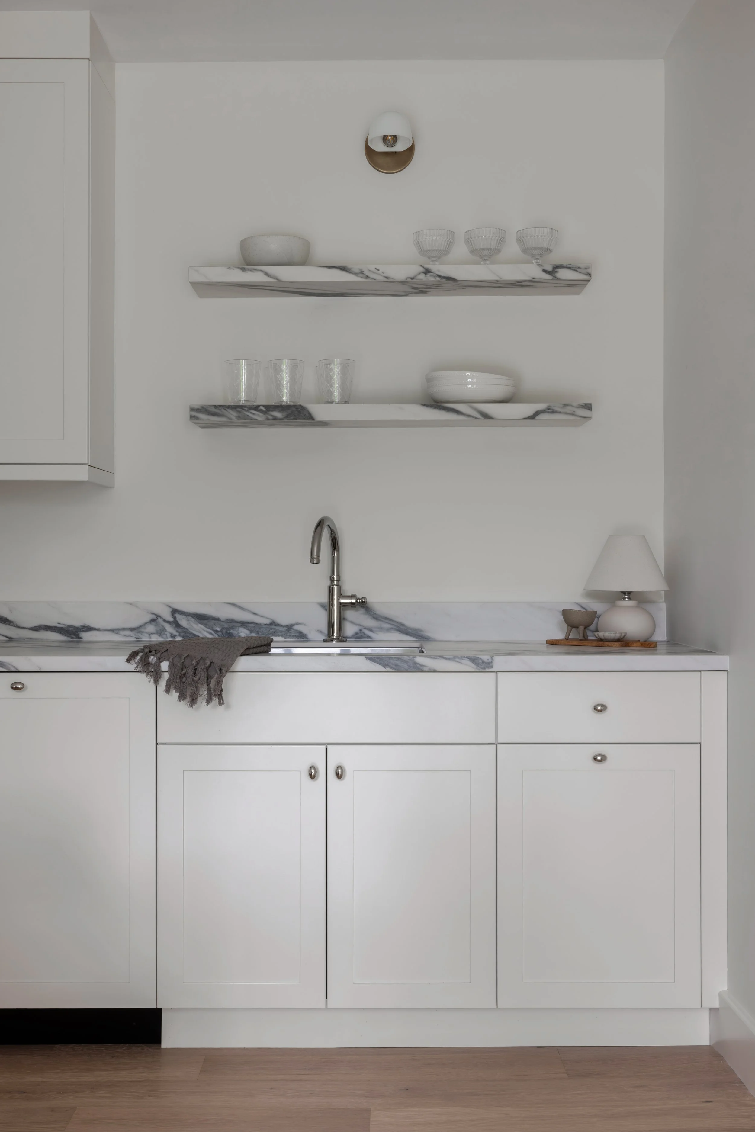A white kitchen cabinet with marble countertops and open shelves displaying bowls, glasses, and dishes. A silver faucet is in the sink, and there is a gray towel on the counter. Light wood flooring completes the scene.