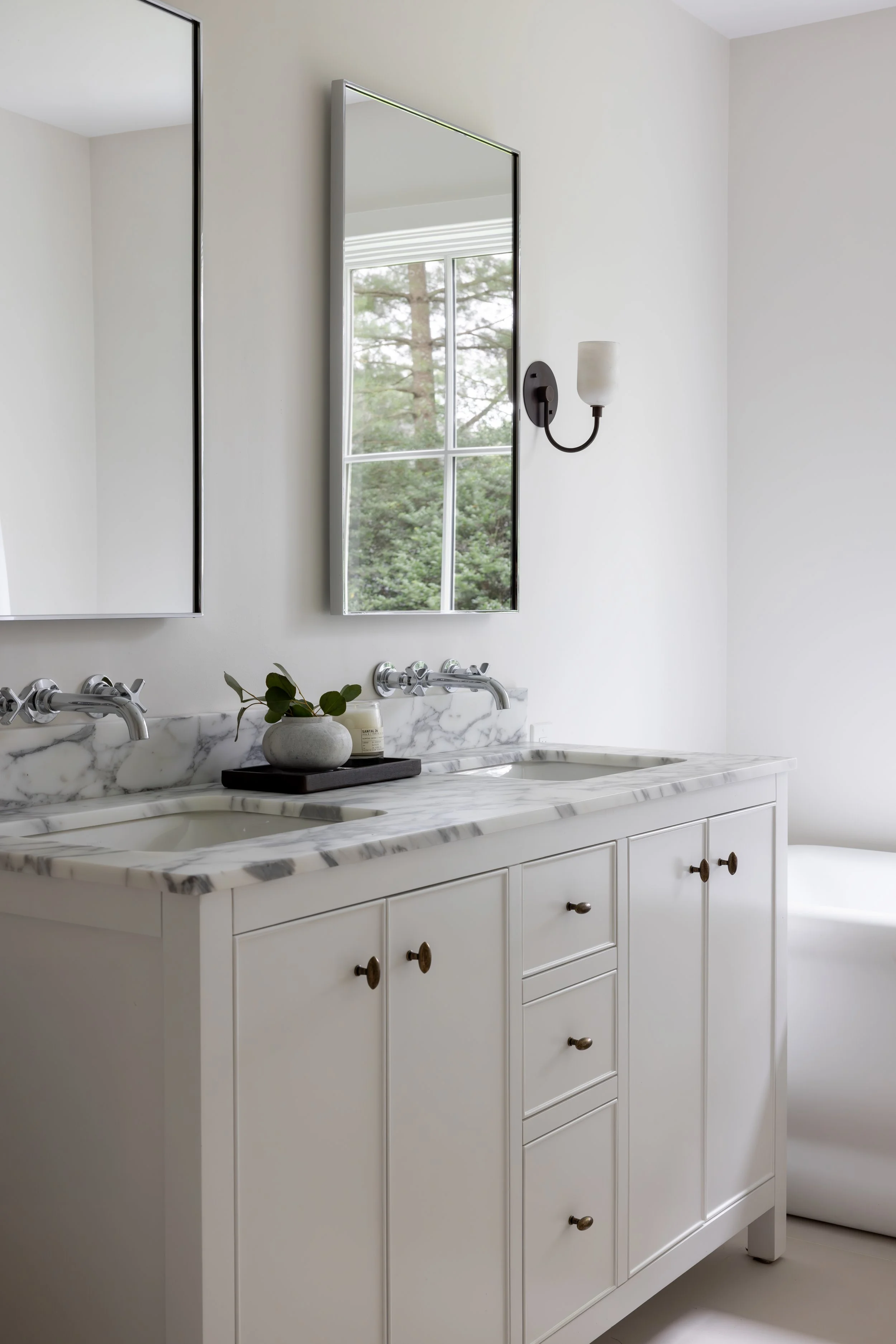 White bathroom vanity with marble countertop, double sinks, and cabinetry, with a mirror, a wall-mounted light fixture, and a window showing trees outside.
