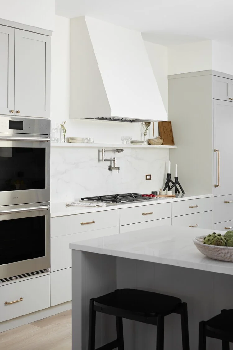 Modern, white kitchen with stainless steel appliances, white cabinetry, and a marble backsplash. Features a stove, a range hood, and decorative items like candles and art on a shelf.