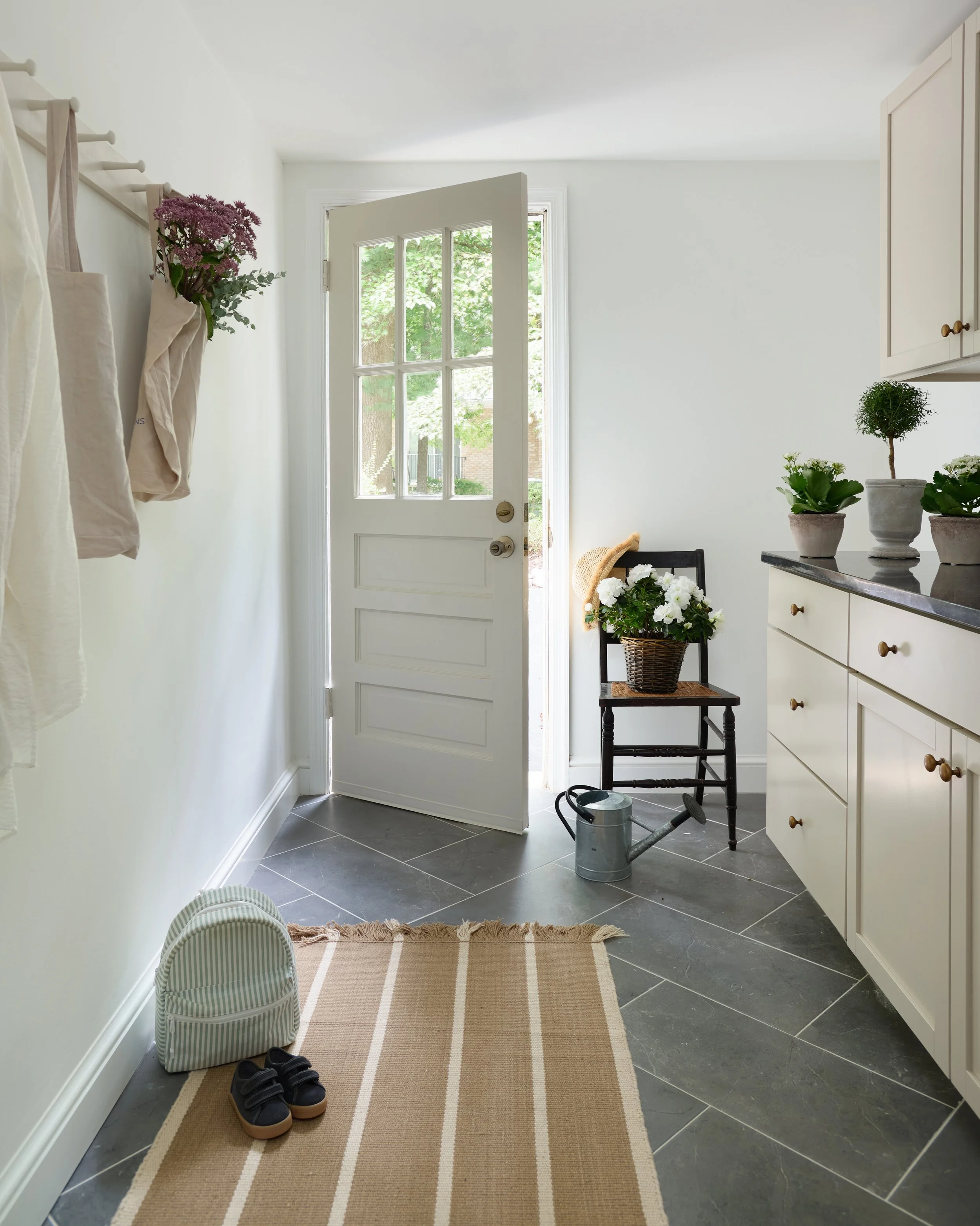 An entryway with a front door open to the outdoors, showing trees and a brick building outside. Inside, there is a beige rug on a dark tiled floor, with a pair of black shoes and a striped bag on the floor, and a small chair holding a basket of white