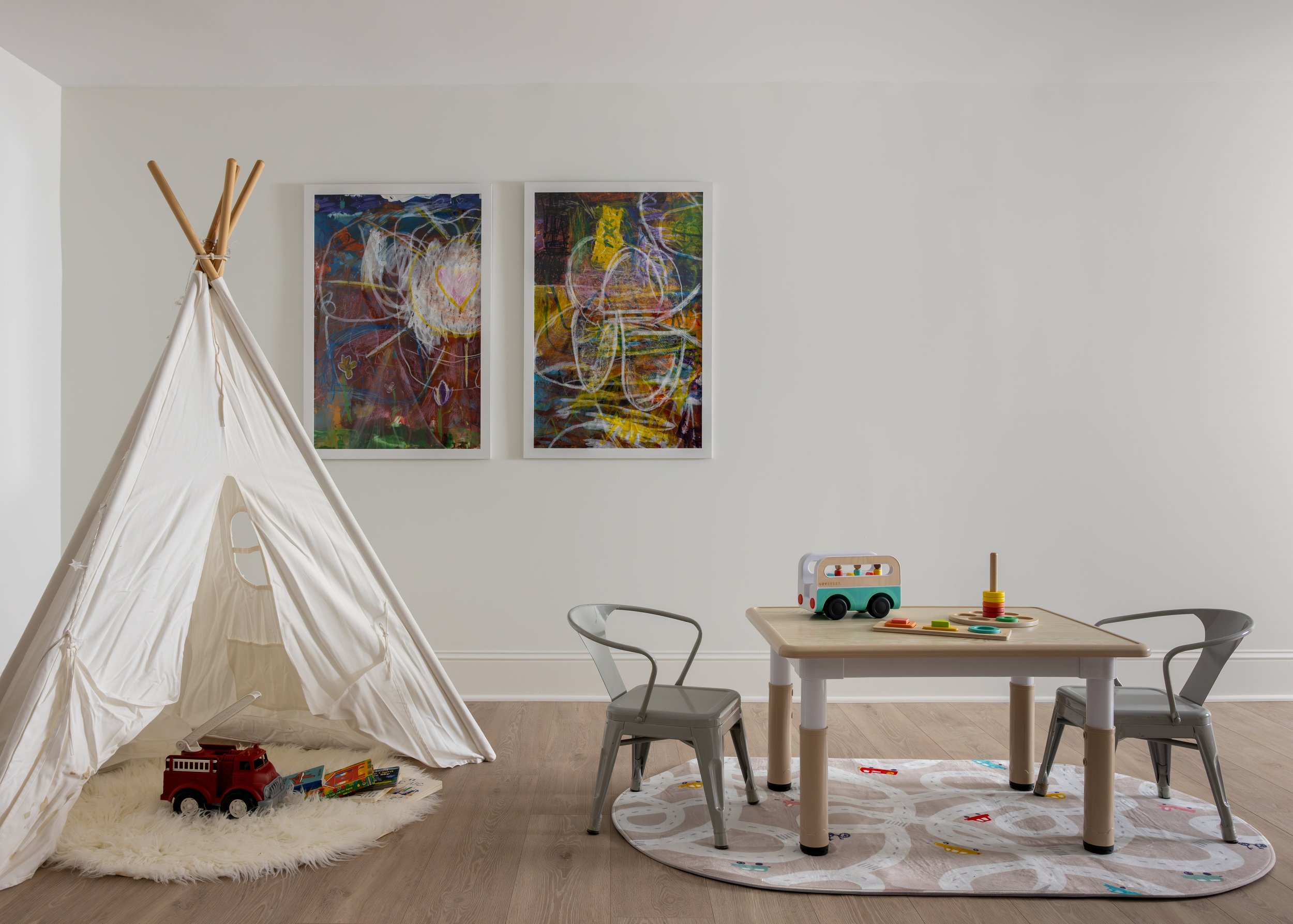 Children's playroom with a small teepee tent, colorful toys including a fire truck and stacking rings on a table, abstract artwork on the white wall, and a circular rug with road patterns.