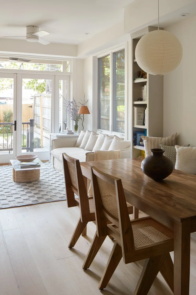 Bright living room with a white sofa, wooden dining table with three chairs, large windows, a paper lantern pendant light, and a sliding glass door leading outside. Decor includes a table lamp, vases, and cushions.