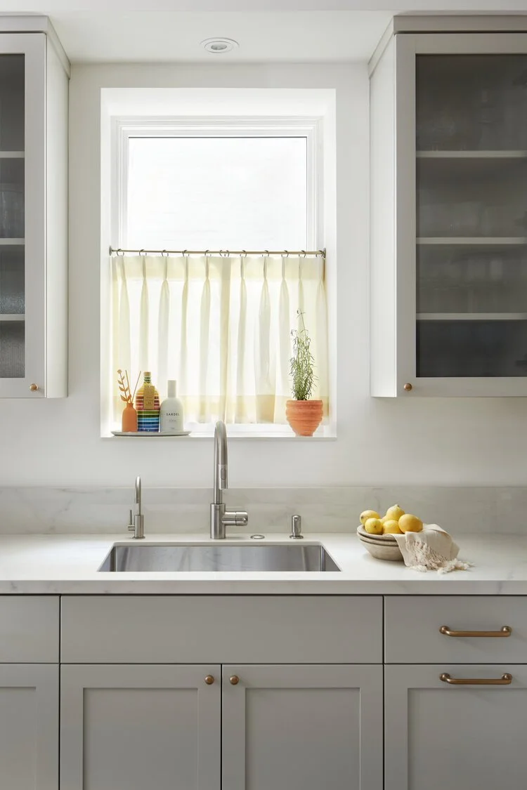 A modern kitchen with white cabinets, a marble countertop, and a stainless steel sink. There is a small window above the sink with a light yellow curtain, and decorative bottles and a potted plant on the window sill. A bowl of lemons and a cloth are 