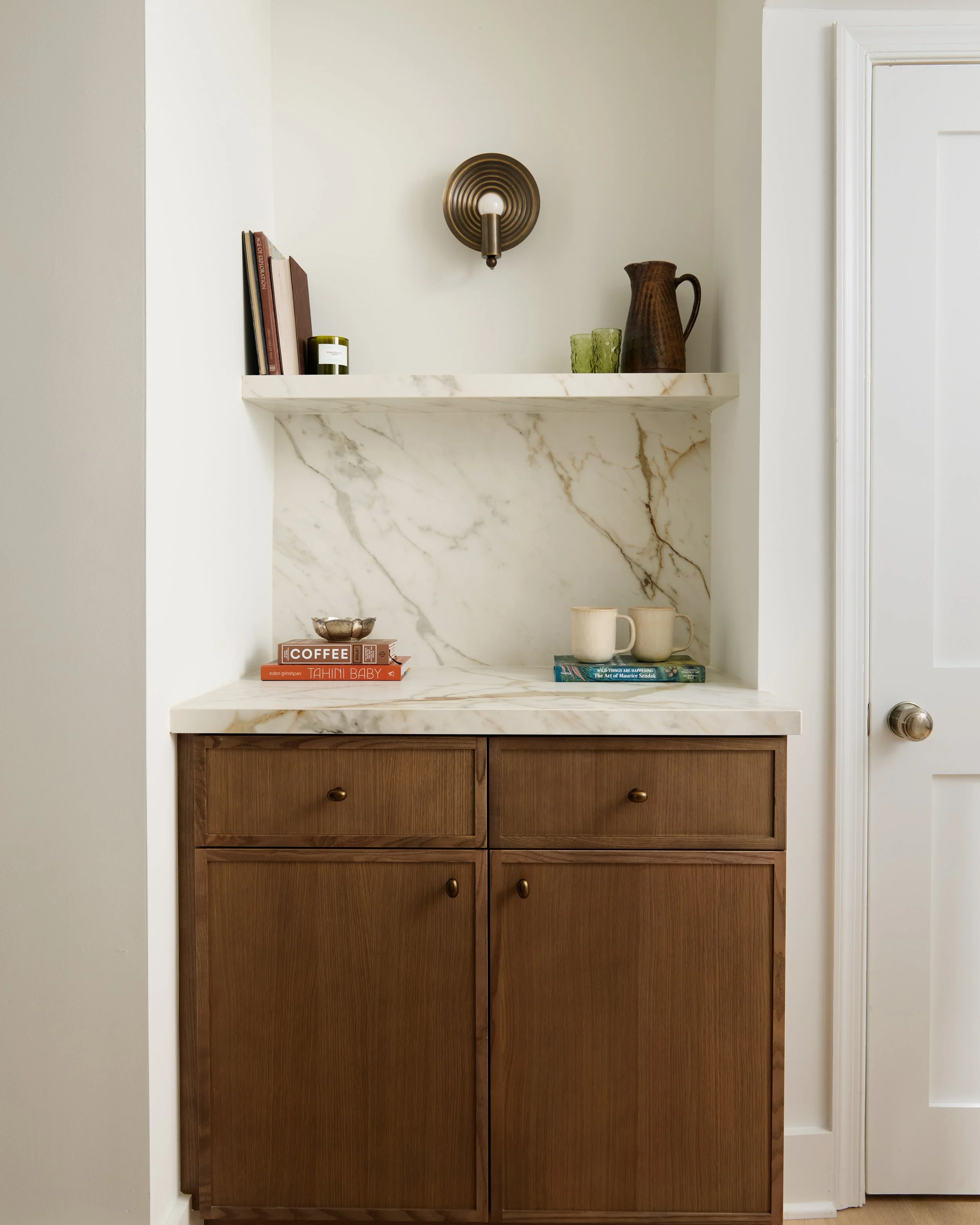 A wooden cabinet with four drawers beneath a marble shelf. The shelf holds books, a candle, a ceramic pitcher, and two green glasses. A brass wall sconce light is mounted above the shelf on the wall.
