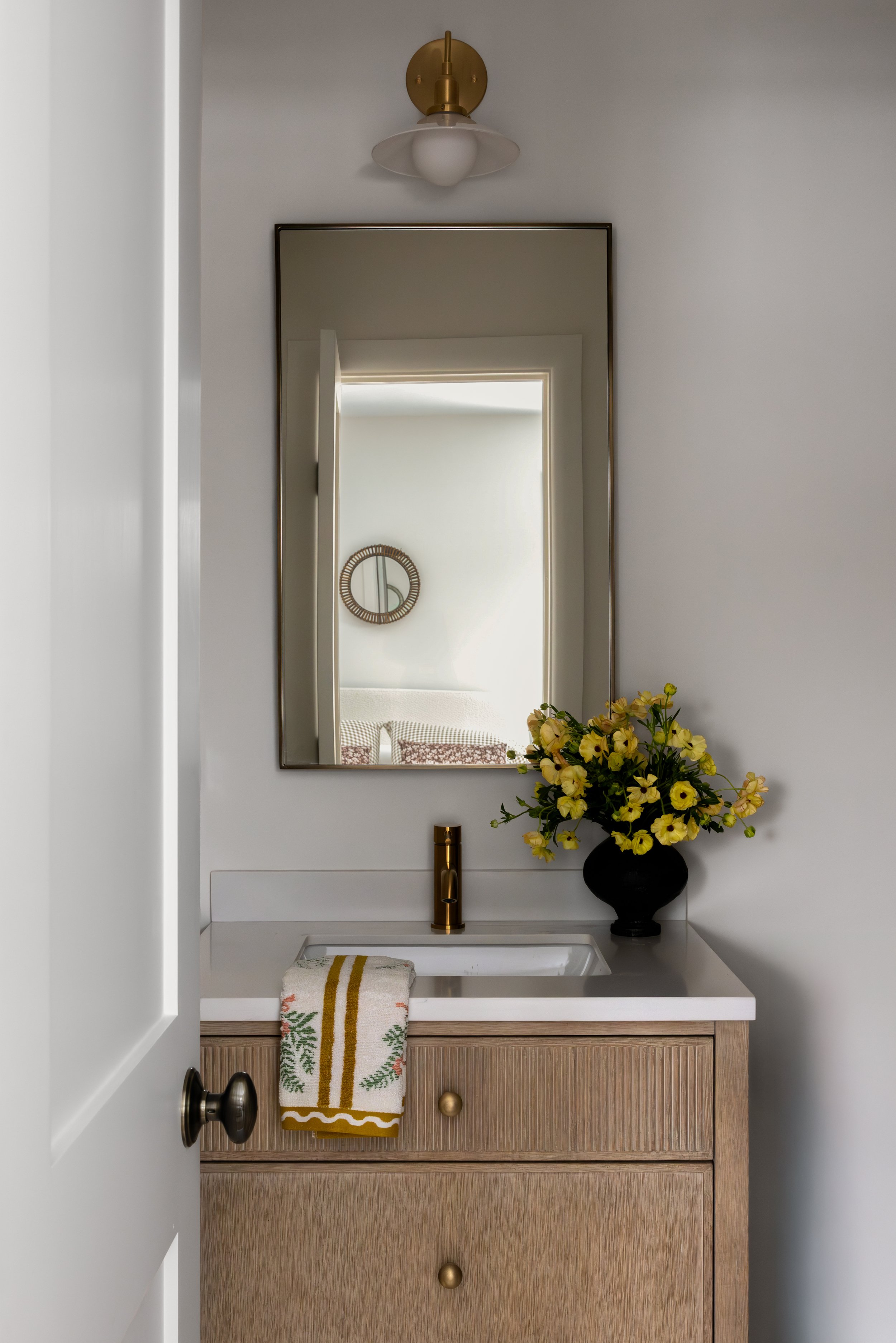 A bathroom vanity with a light wood cabinet, a white countertop with an integrated sink, and a gold faucet. A black vase with yellow flowers sits on the counter, with a gold and white towel hanging on the side. A mirror, a wall-mounted light fixture,
