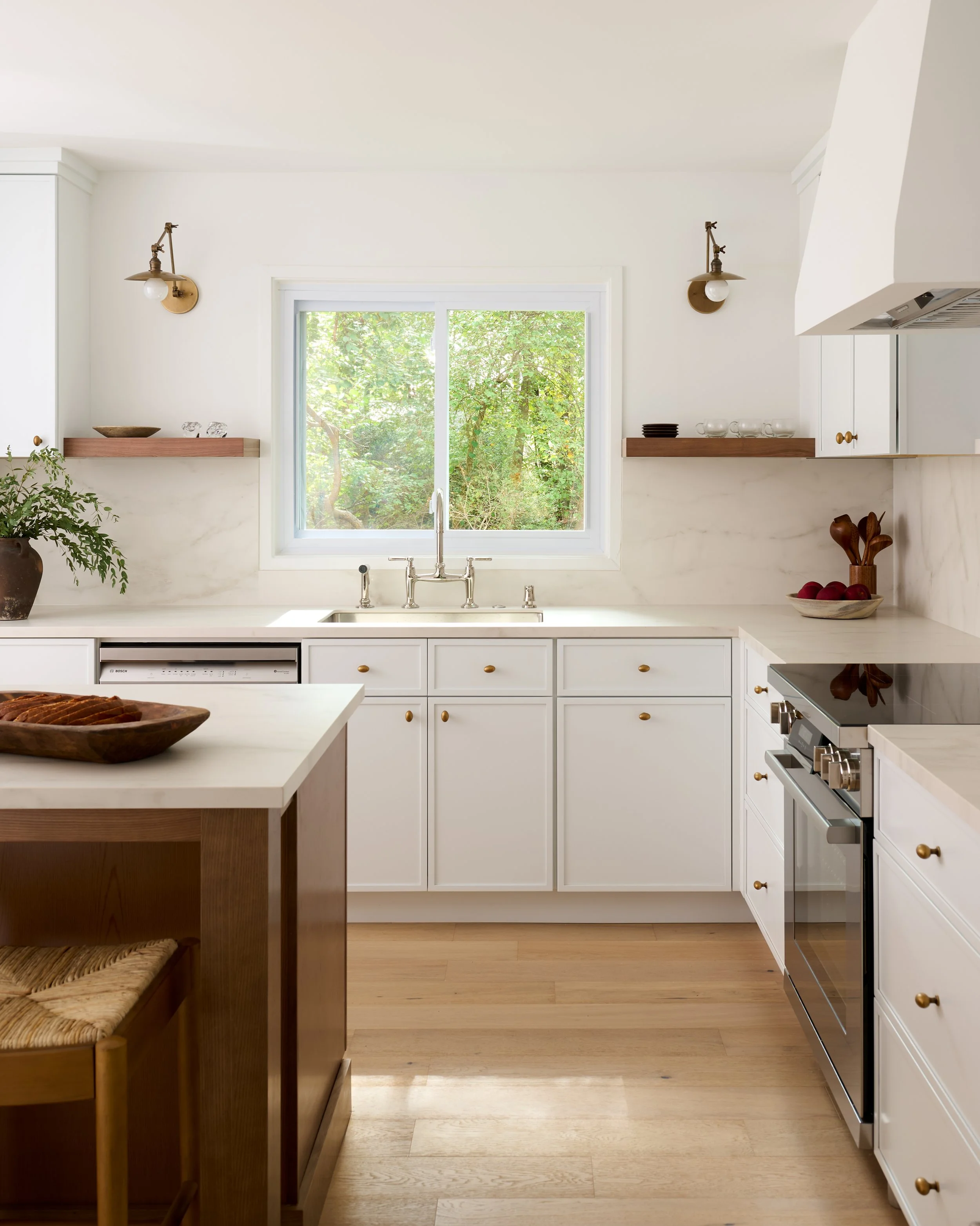 A modern white kitchen with a large window showing greenery outside. The kitchen has white cabinetry, brass handles, and wooden open shelves with dishes and decorative items. There is a wooden island with a bowl of bread on it and a potted plant on t