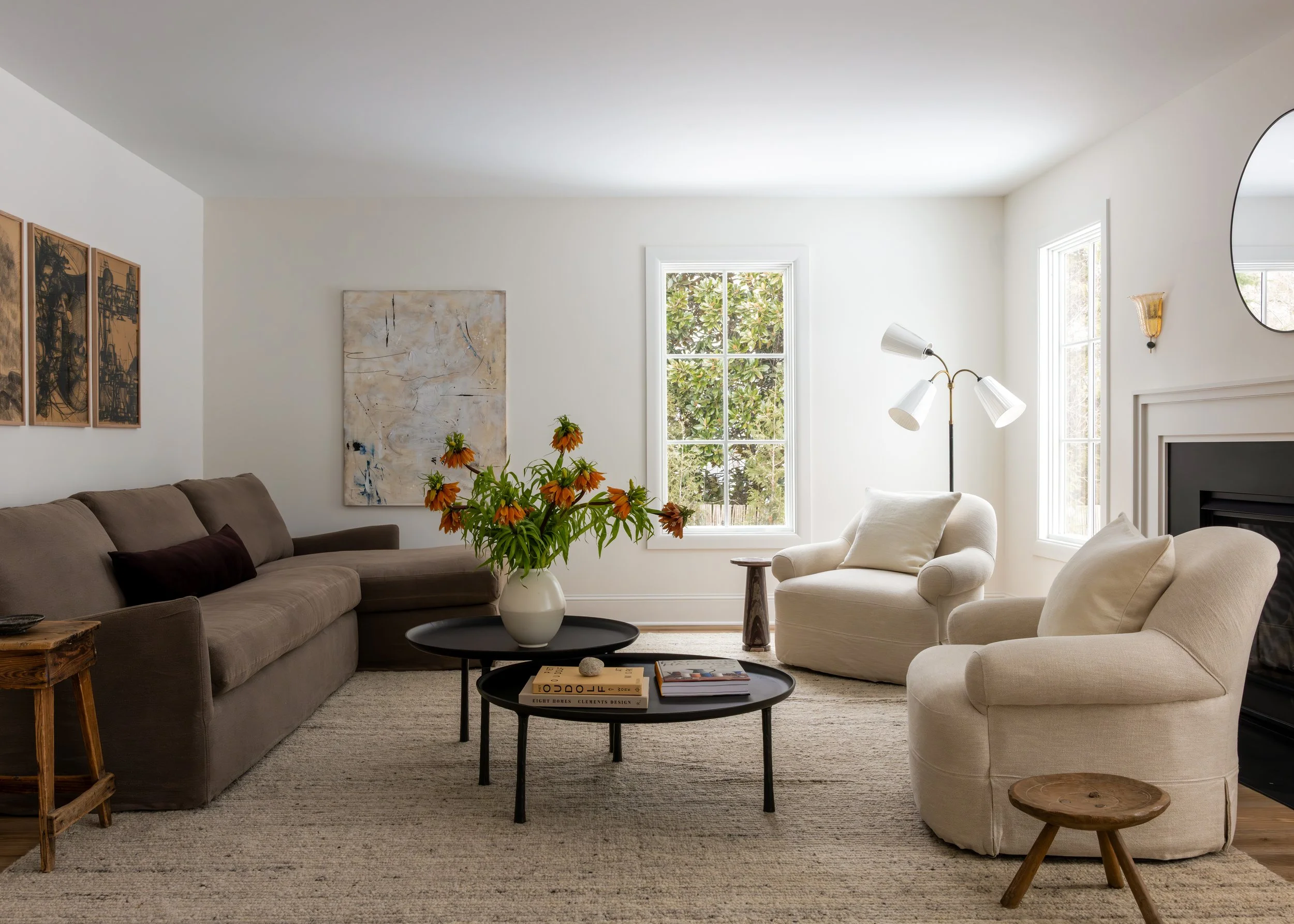 Bright living room with large windows, a beige sofa with dark cushions, white armchair, black coffee tables with books, a vase of orange flowers, wall art, floor lamp, and wall-mounted sconce.