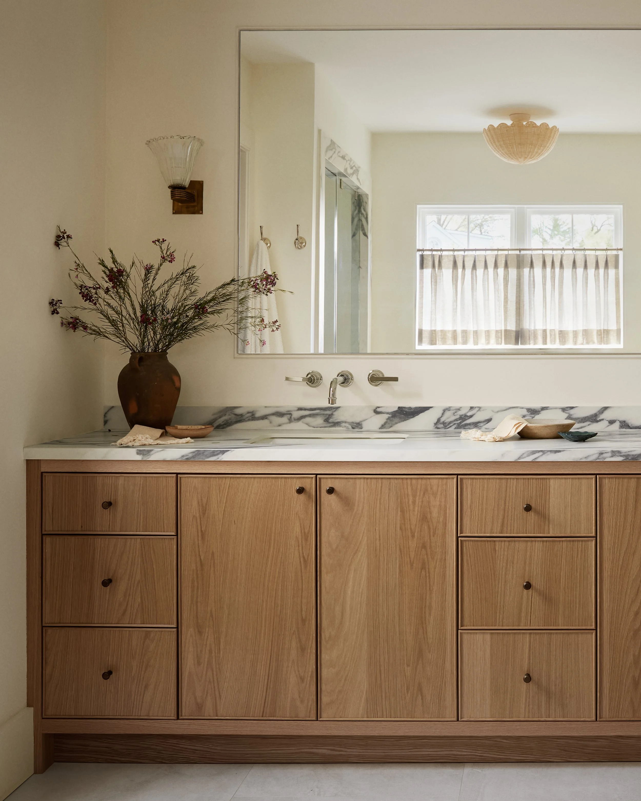 A bathroom vanity with a marble top, wooden cabinetry, and a large mirror. A brown vase with dried flowers sits on the left side of the counter. There are three round knobs on the drawers and two full-length handles on the cabinet doors. A window wit