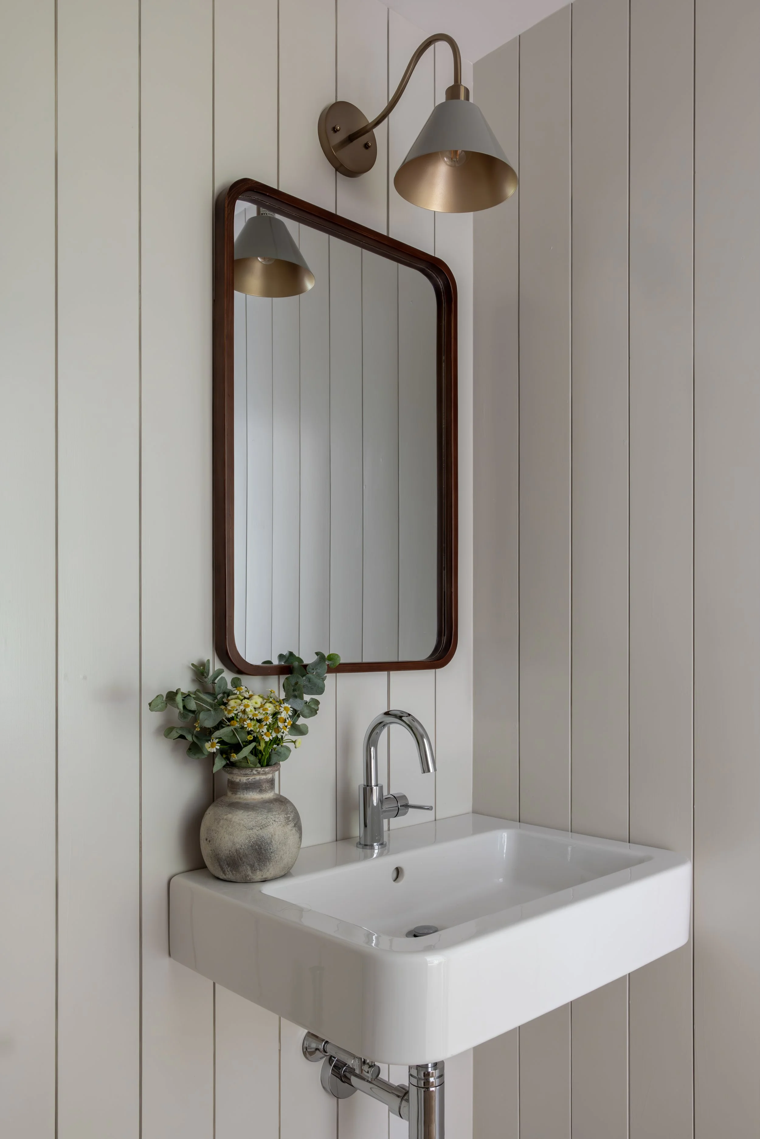 Bathroom with wall-mounted sink, faucet, framed mirror, wall sconce, and a vase with flowers on the sink counter.