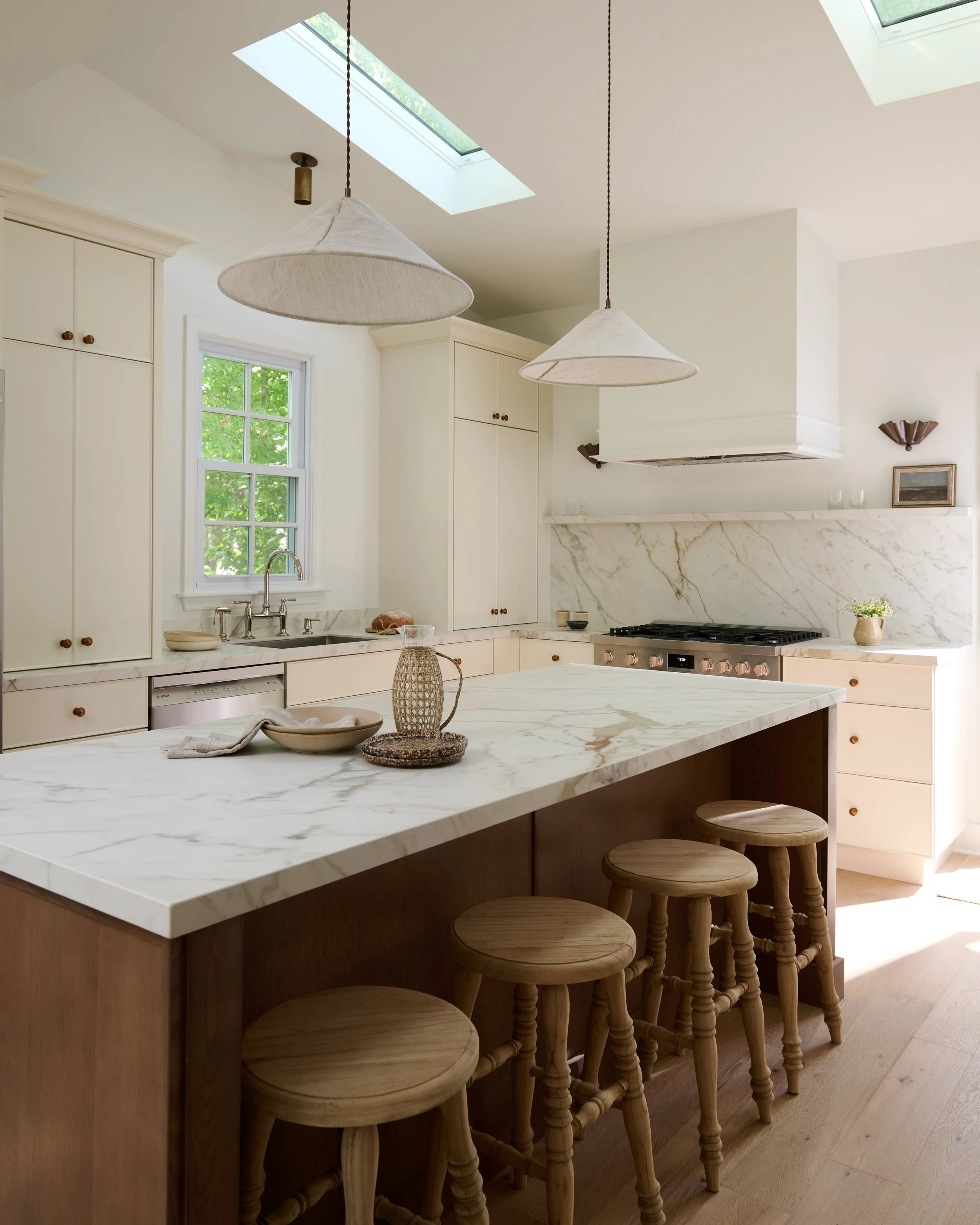 A modern kitchen with white cabinets, a marble countertop, and a wooden island with five wooden stools. Two pendant lights hang over the island, and there are skylights and a window with greenery outside.