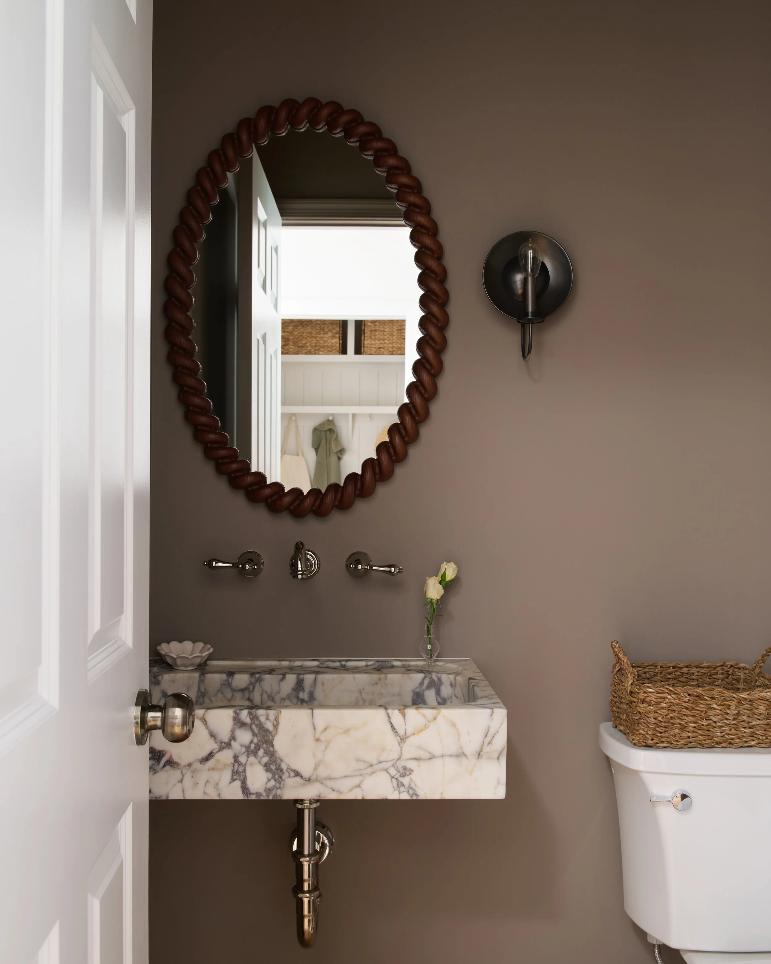A bathroom with a marble sink, a mirror with a brown frame, a wall-mounted light, a small vase with white roses, and a woven basket on a white toilet tank.