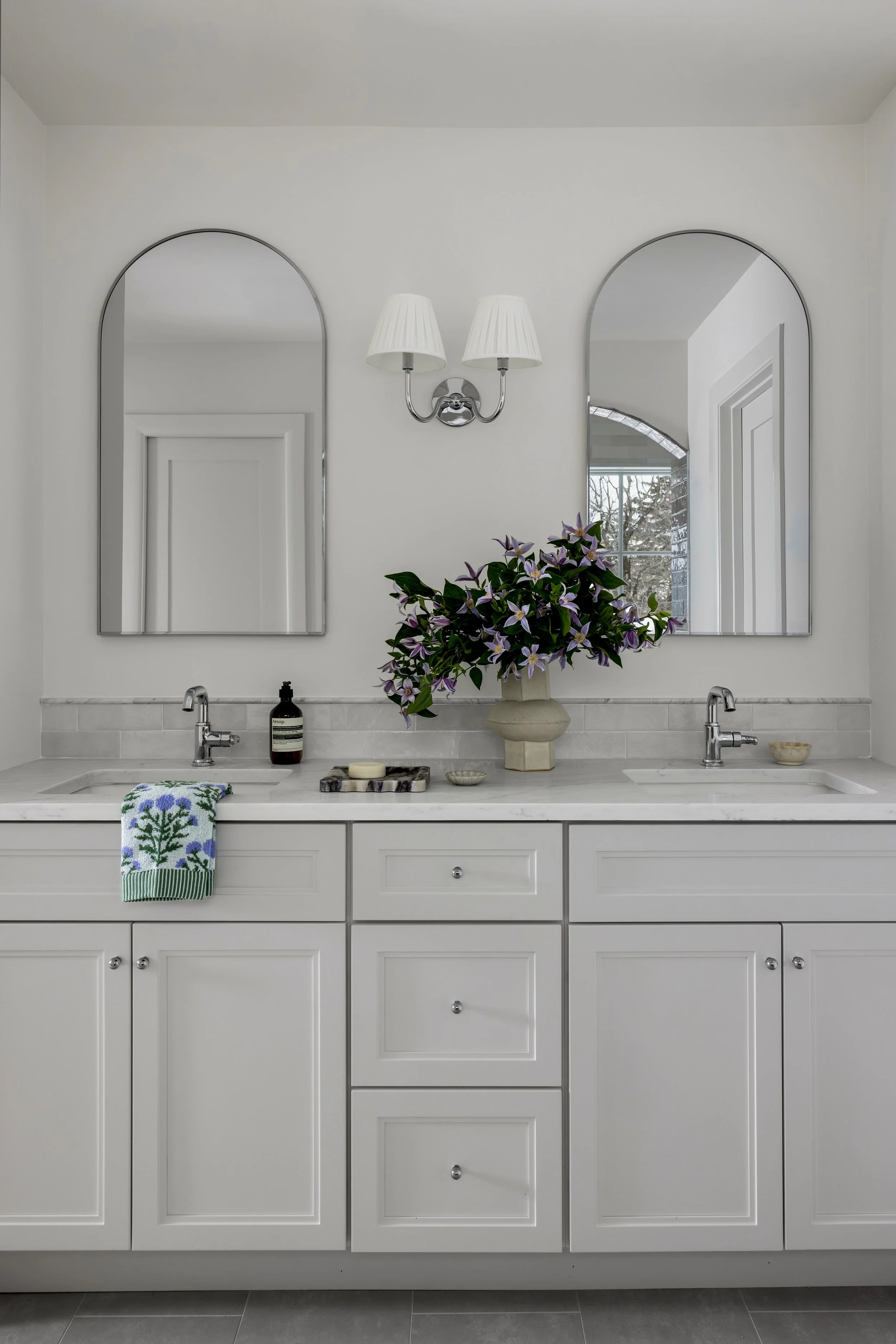 A bathroom vanity with a white marble countertop, white cabinets, and two silver sinks with silver faucets. Two rectangular mirrors with rounded tops are mounted above the sinks and a wall-mounted double-shade wall sconce is in between. A purple flow