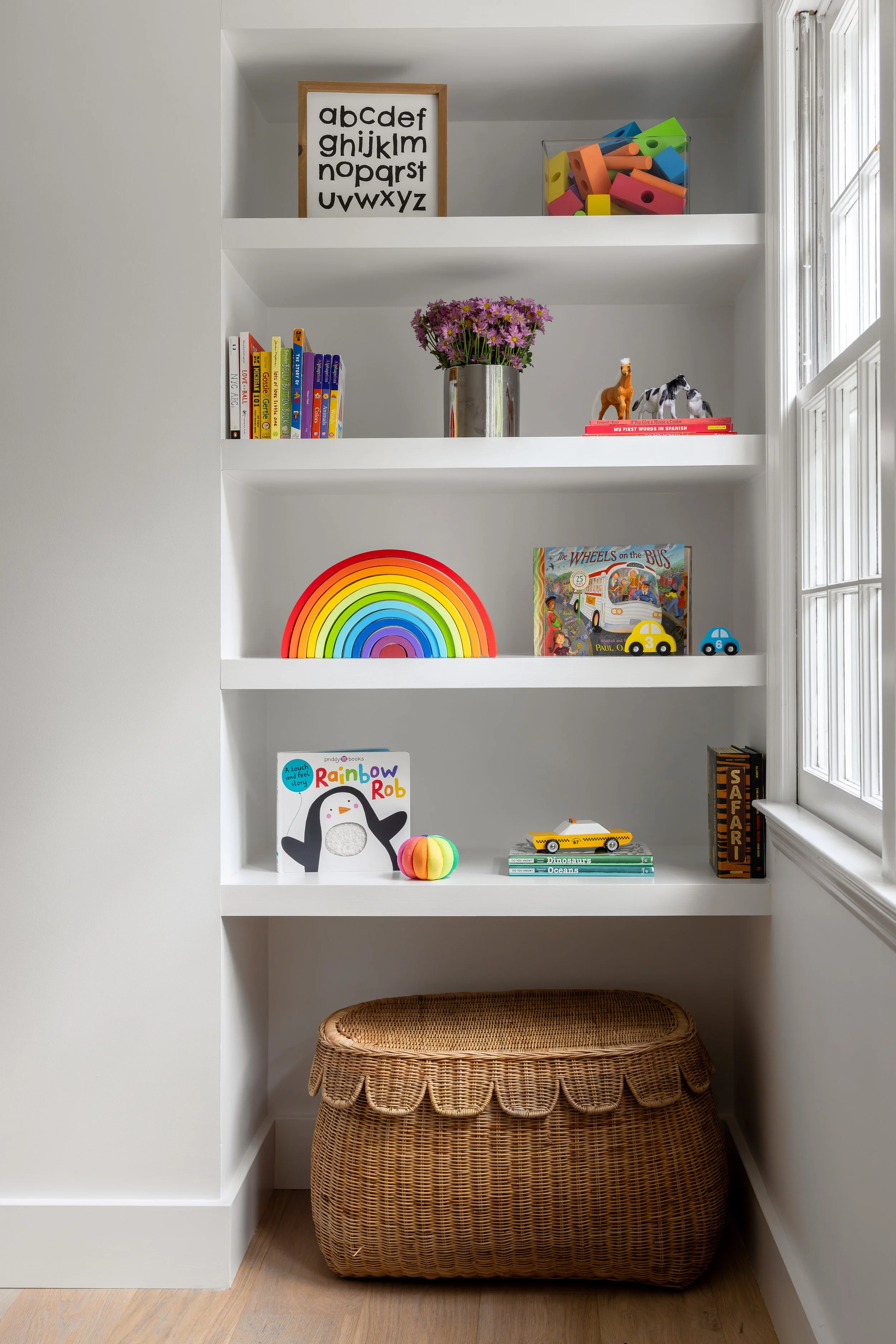 White built-in bookshelf with four shelves filled with children's books, toys, and decor. The top shelf has a framed alphabet print and colorful foam blocks. The middle shelf has a vase of flowers, toy animals, and books. The third shelf has a rainbo