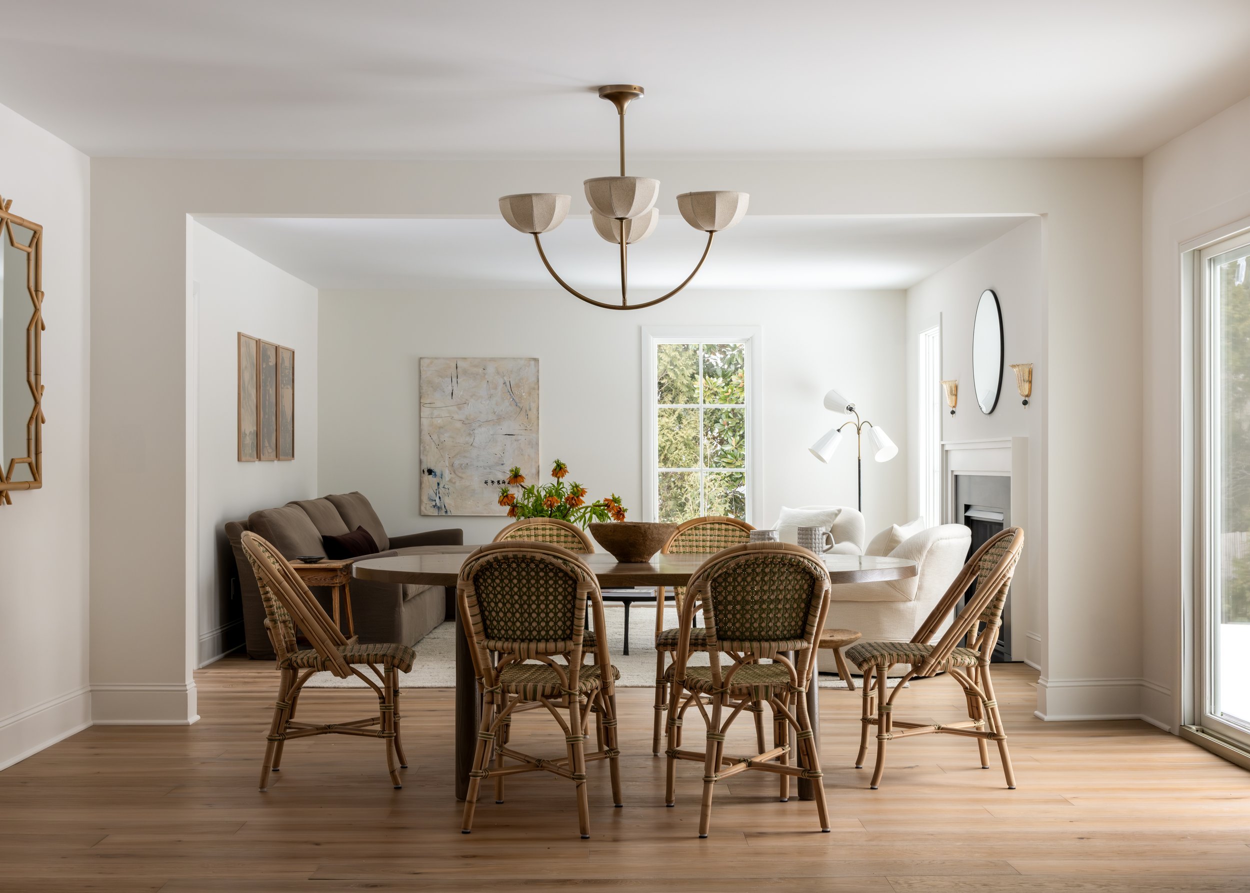 A bright, airy living space with a round wooden dining table surrounded by six wicker chairs, a beige sofa, a white armchair, a large window, artwork on the wall, and a modern chandelier hanging from the ceiling.