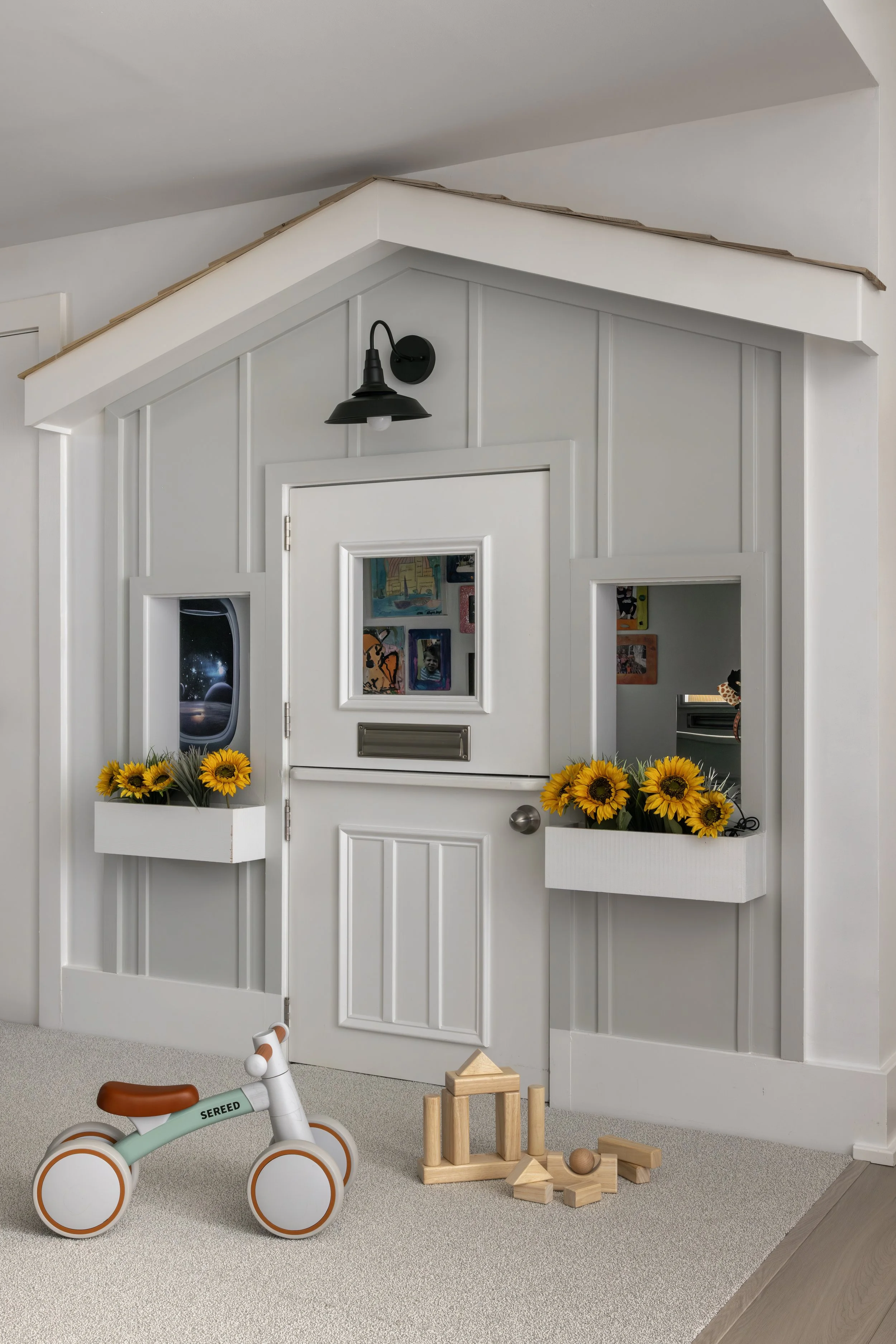Children's play area with a white handcrafted wall, small window displaying space-themed artwork on the wall, a black wall-mounted light, artificial sunflowers in white planters, a wooden toy block set on beige carpet, and a wooden floor transition.
