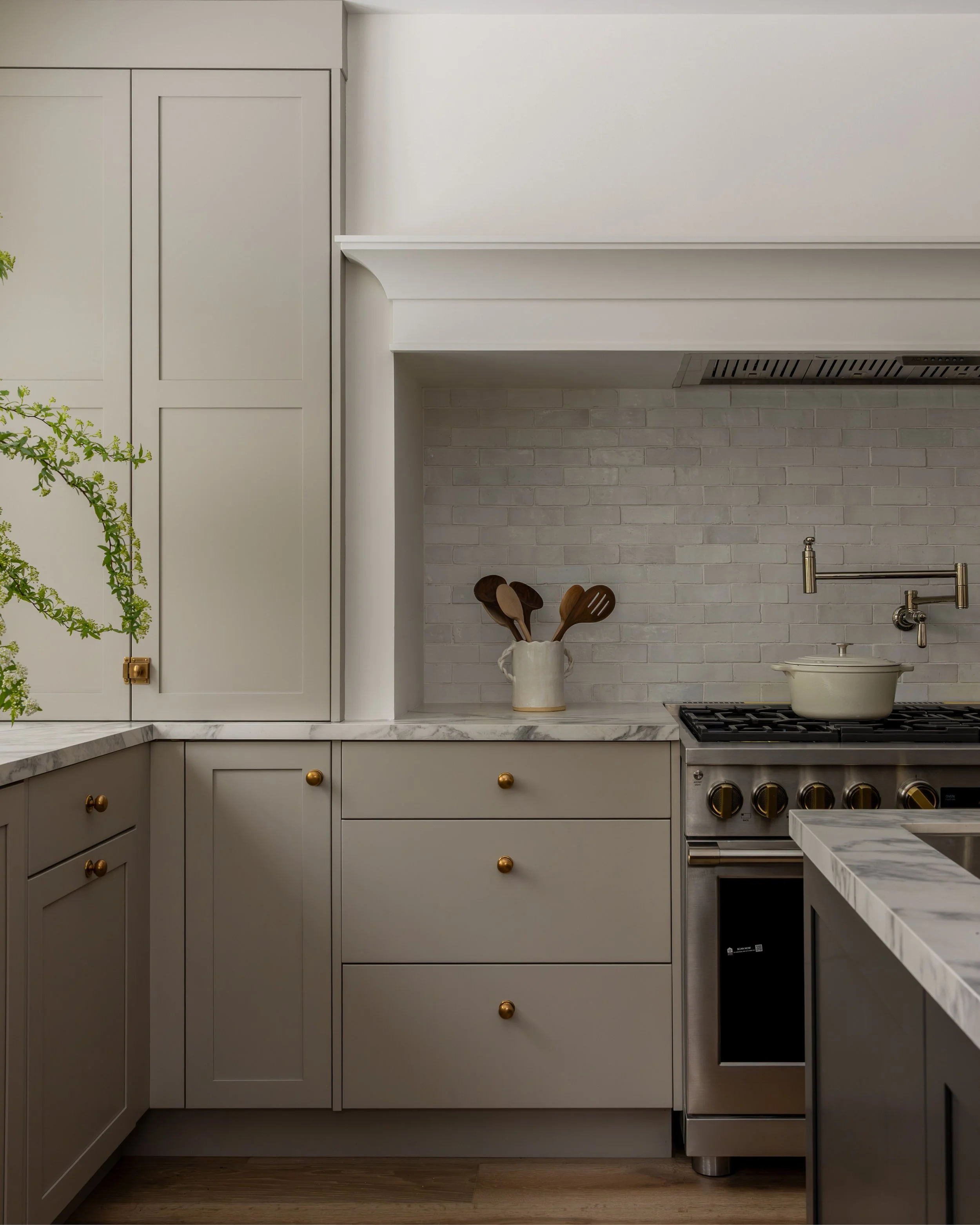 Kitchen with white cabinets, marble countertops, a white brick backsplash, a ceramic utensil holder with wooden spoons, and a stove with a pot, under a white range hood.