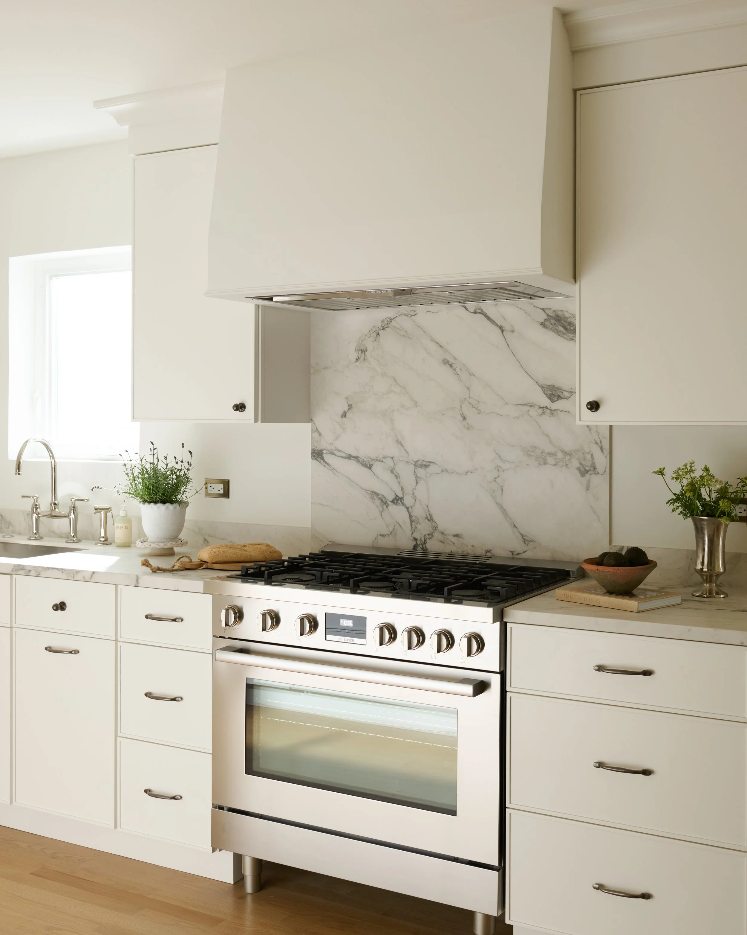 Modern kitchen with white cabinets, marble backsplash, and a stainless steel oven and range. There are small plants on the countertop and a window letting in natural light.