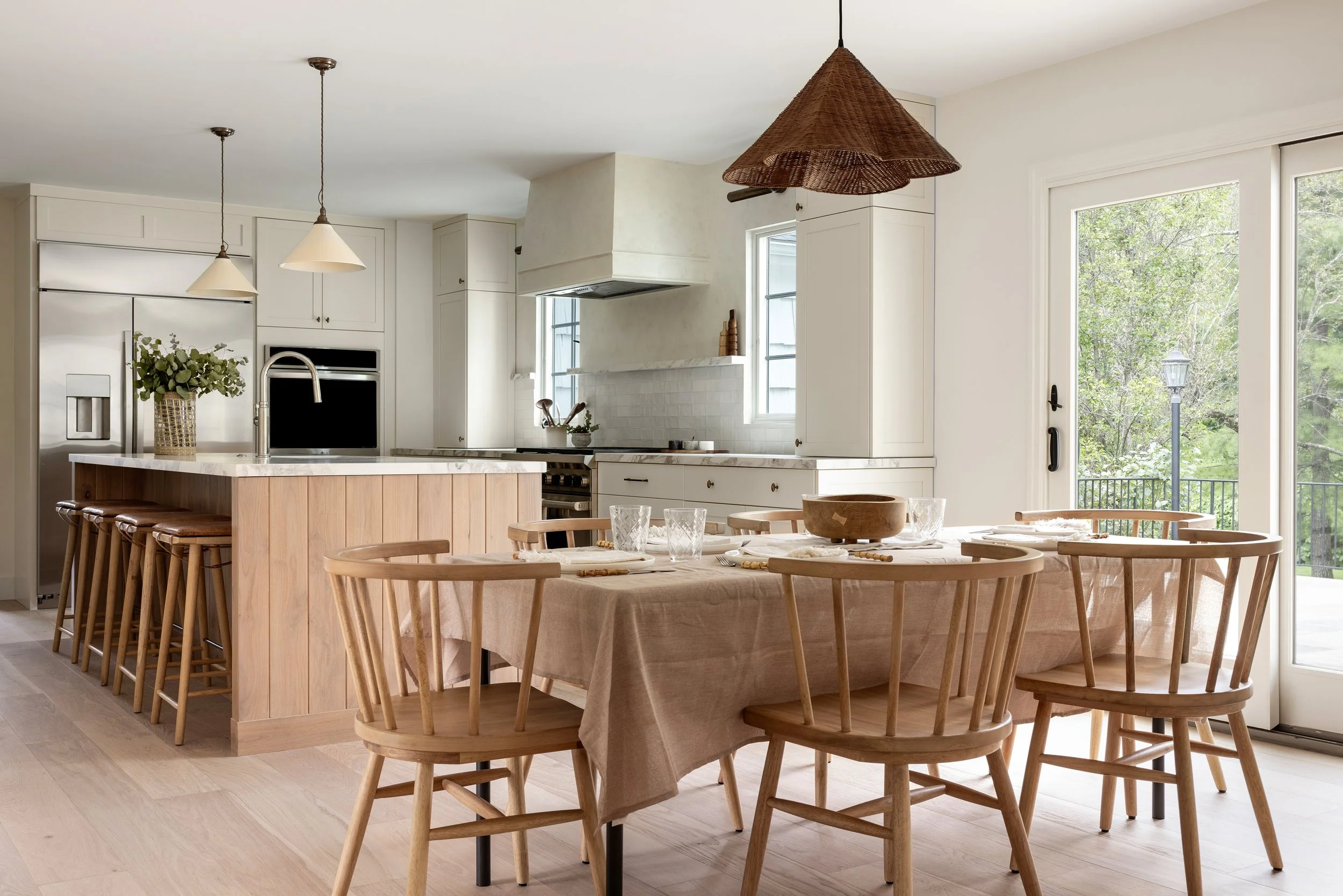 Modern kitchen with white cabinets, stainless steel appliances, and a wooden dining table with matching chairs, overlooking a green outdoor space through large sliding glass doors.
