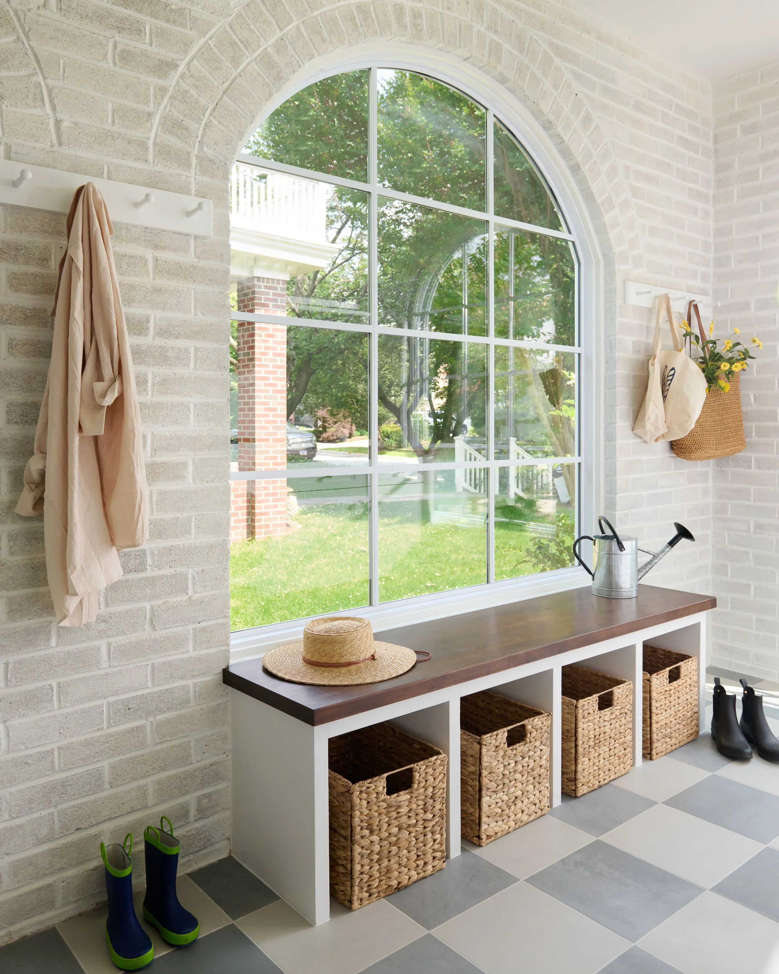 Entryway with a window, white brick walls, storage bench with baskets, a hat, watering can, and rain boots.