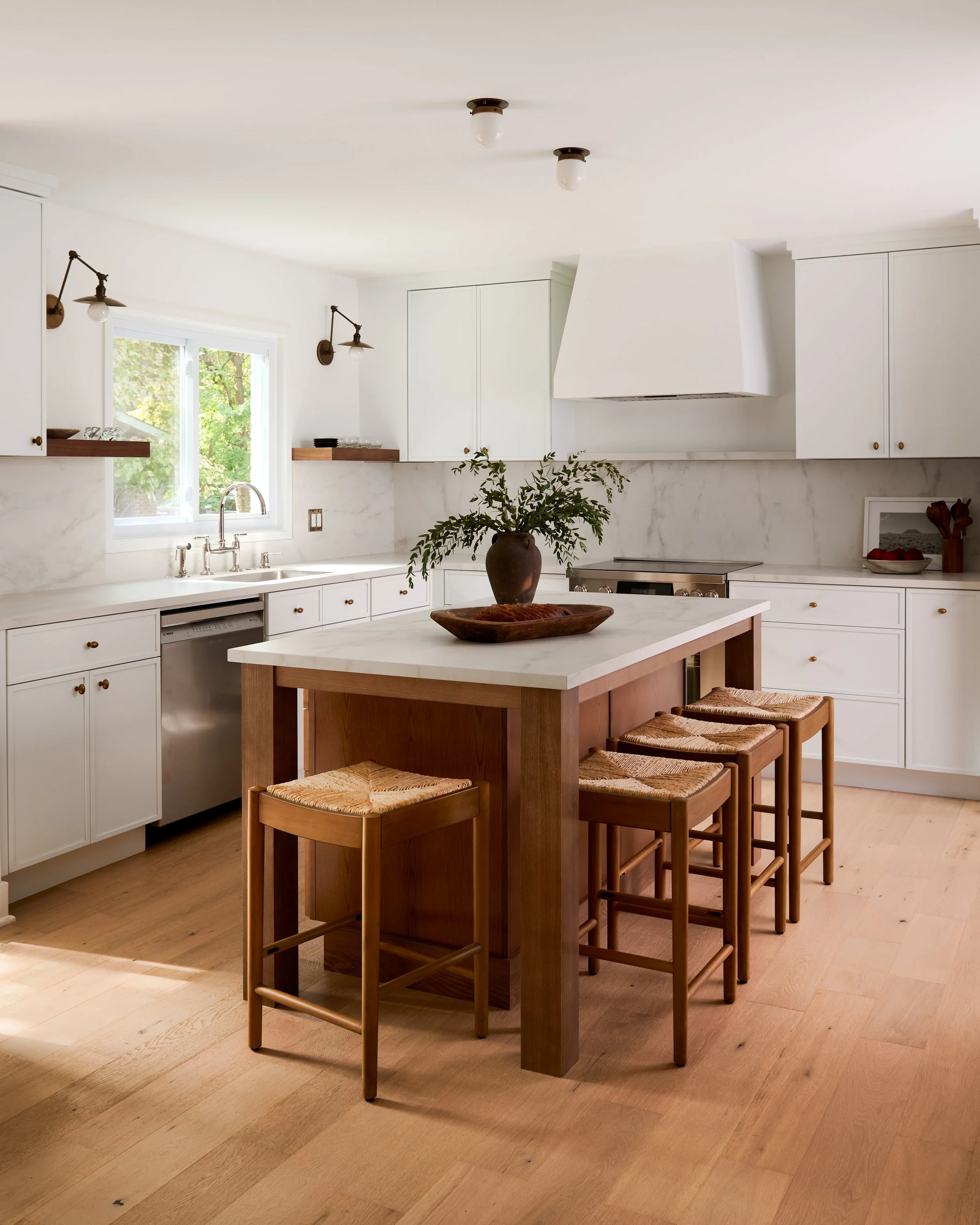 A modern kitchen with white cabinets, wooden stools around a wooden island, and a window showing trees outside.