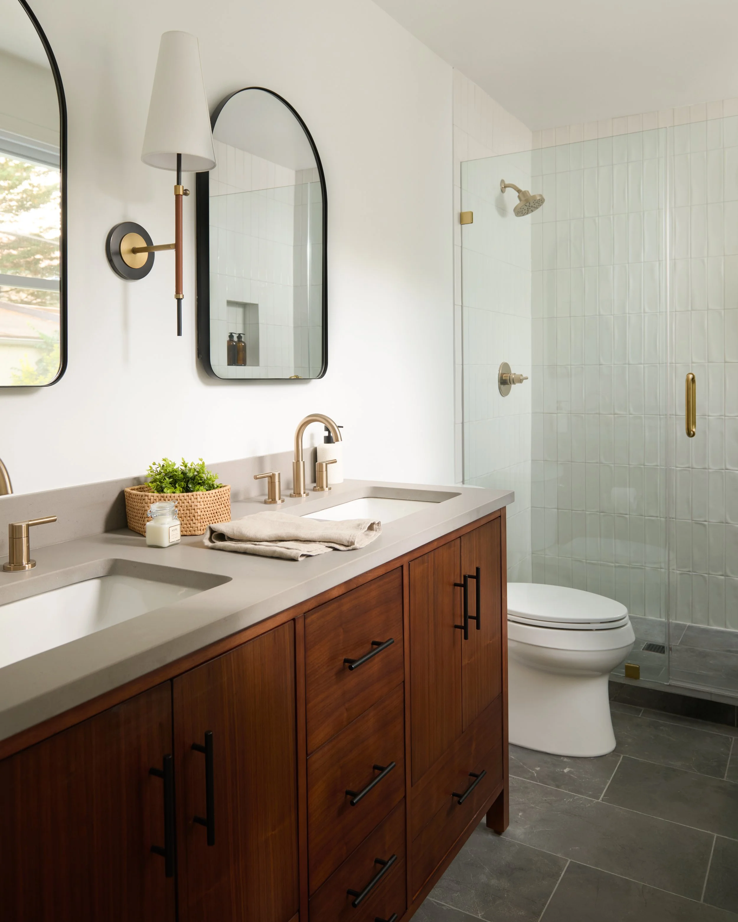 A modern bathroom featuring a wooden vanity with a white countertop, a sink, a gold faucet, and a decorative plant. Two mirrors are mounted on the white wall, and the bathroom includes a glass-enclosed shower with white tile walls, a showerhead, and 