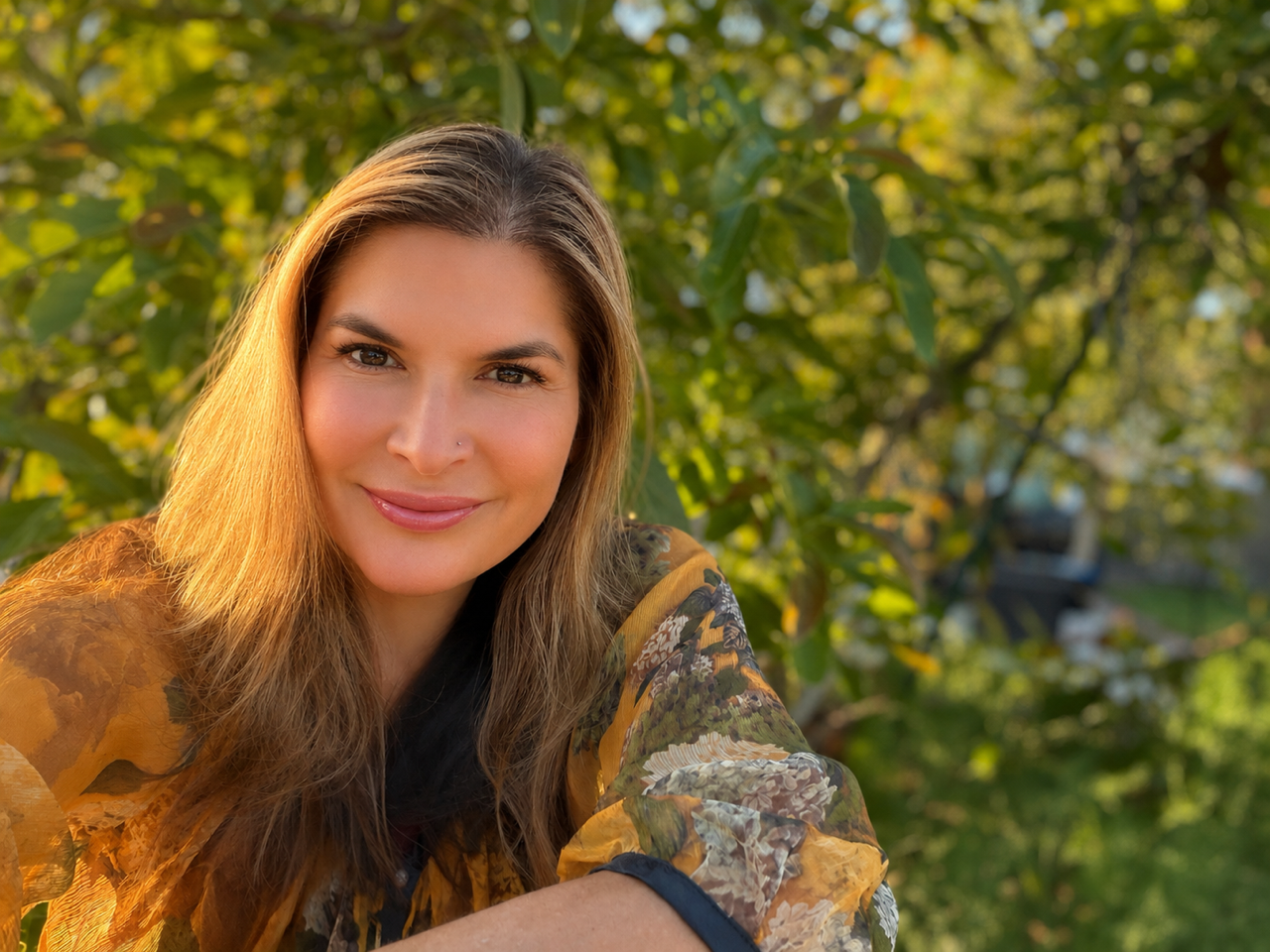 A woman with long brown hair and a nose piercing smiling outdoors in front of green foliage.