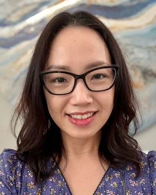 A woman with shoulder-length dark hair, wearing glasses and a blue patterned blouse, smiling in front of a textured background.