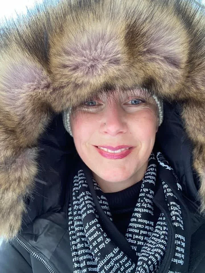 Person with light skin, blonde hair, wearing a fur hat and black jacket, smiling outdoors.