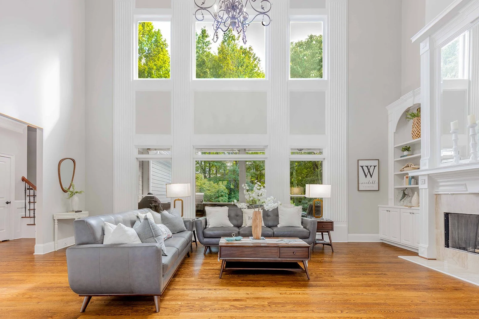 Bright living room with tall windows, white walls, gray sofas with white and gray pillows, wooden coffee table with decorative items, and a white built-in fireplace with candles and decor.