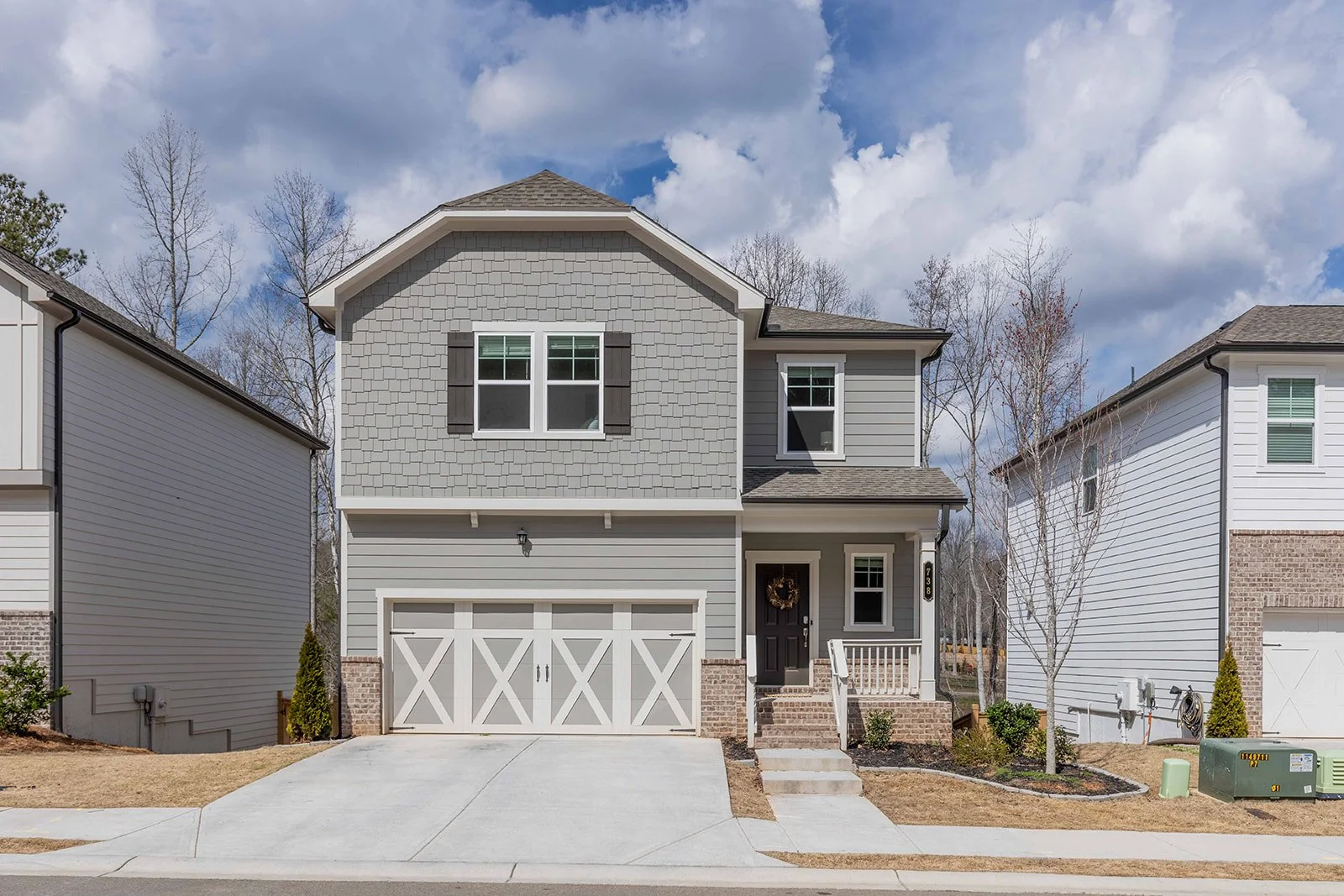 A two-story modern house with gray siding, black shutters, and a front porch. The front yard has a concrete driveway, small trees, and landscaping, with neighboring houses on each side.