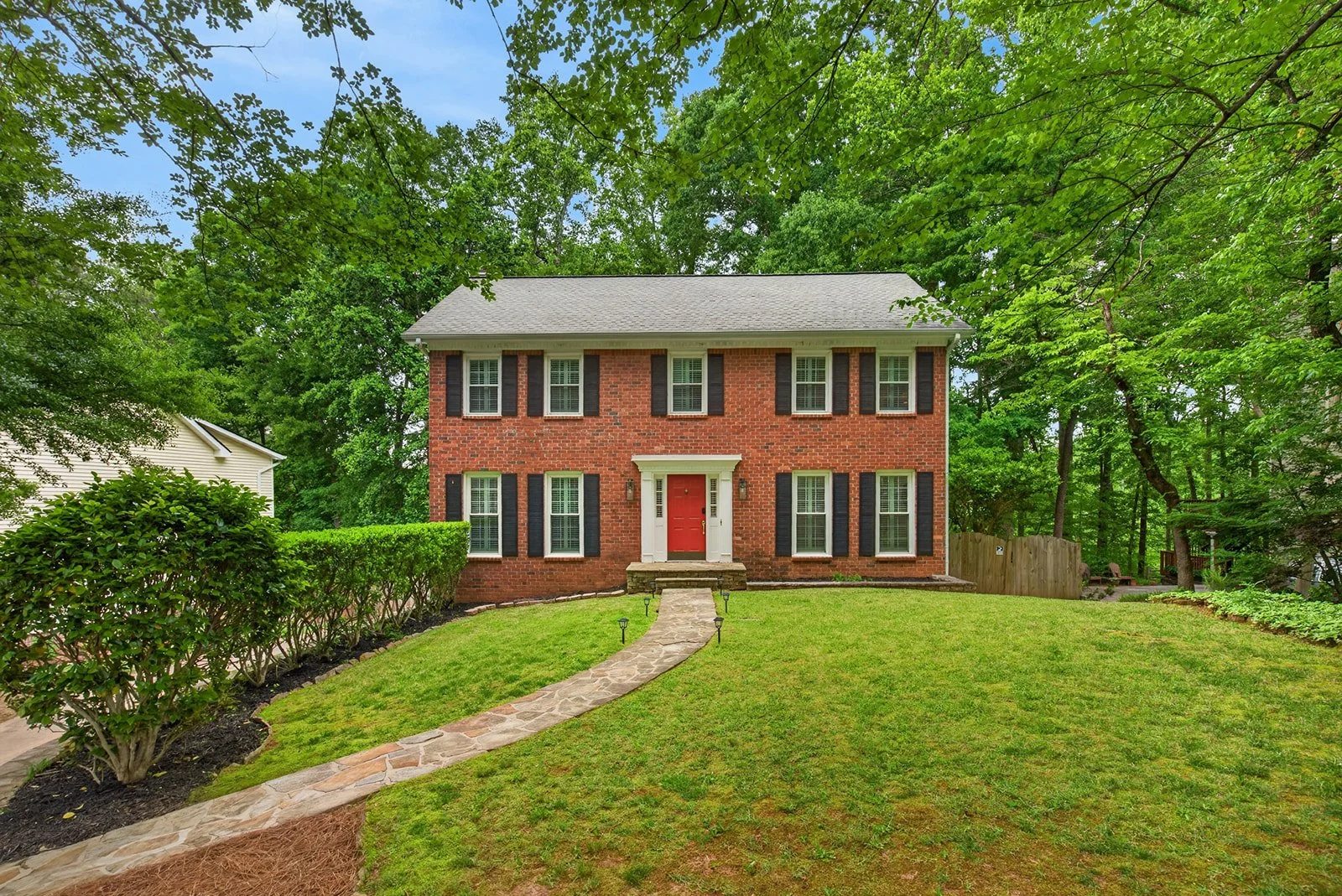 Front view of a two-story brick house with a red front door, black shutters, and a stone pathway leading to the entrance, surrounded by green grass, shrubs, and tall trees.