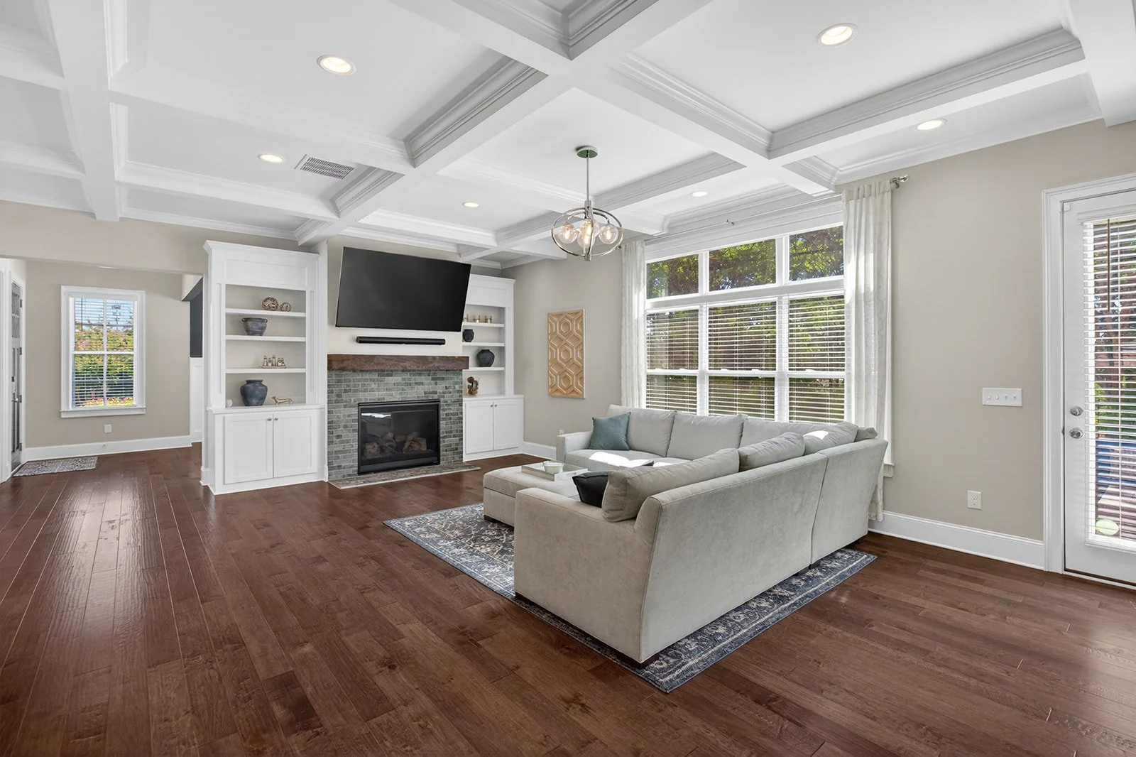 Bright living room with large windows, white walls, hardwood floors, a white sectional sofa, a gray area rug, a fireplace with a brick surround, and a wall-mounted TV above the fireplace. White built-in shelves flank the fireplace, and a modern chand