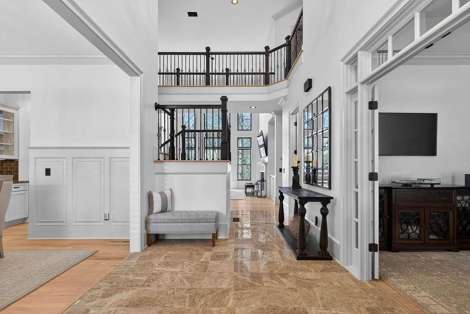 Interior view of a spacious, modern home with white walls, wooden flooring, and black railing staircase leading to an upper level. There is a gray upholstered bench with a pillow, a dark wooden console table with candles, and a large flat-screen TV o