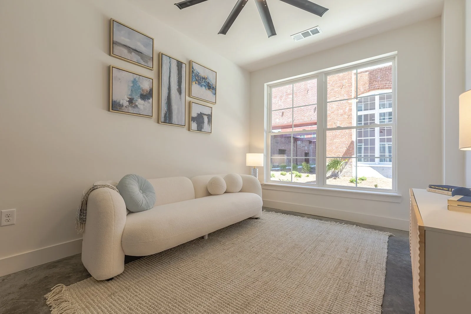 A minimalist living room with a white sofa, abstract artwork on the wall, a large window letting in natural light, a rug on the floor, and a side table with books and a lamp.