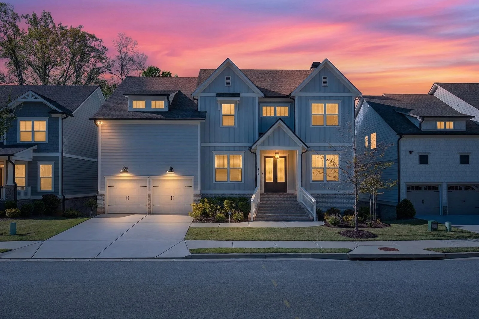 A modern two-story house at sunset with warm interior lights, a driveway, a front porch, landscaped yard, and neighboring houses, under a colorful sky.