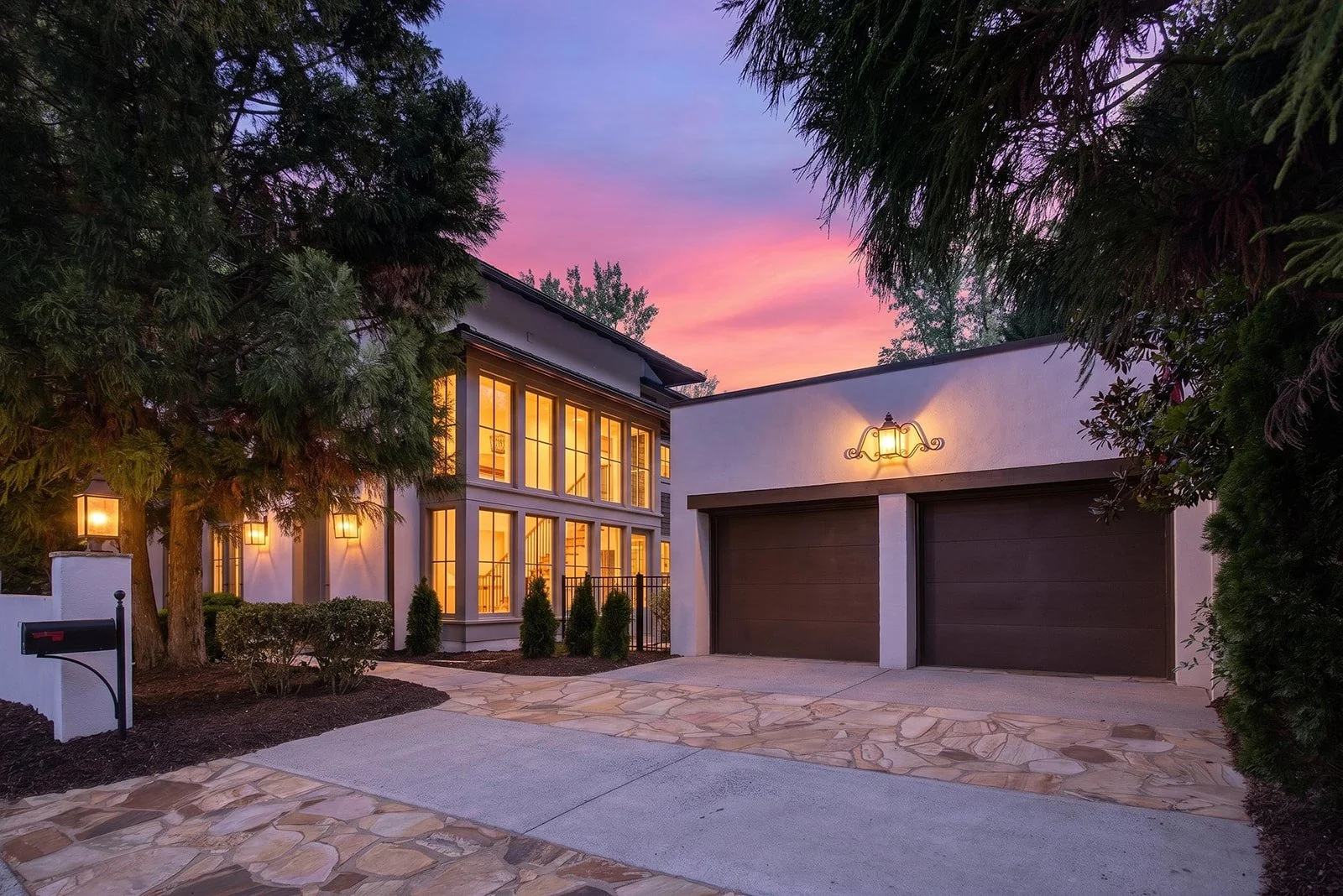 Front view of a modern house at sunset, with lit windows and exterior lights, surrounded by trees, a stone driveway, and a garage with two dark-colored doors.