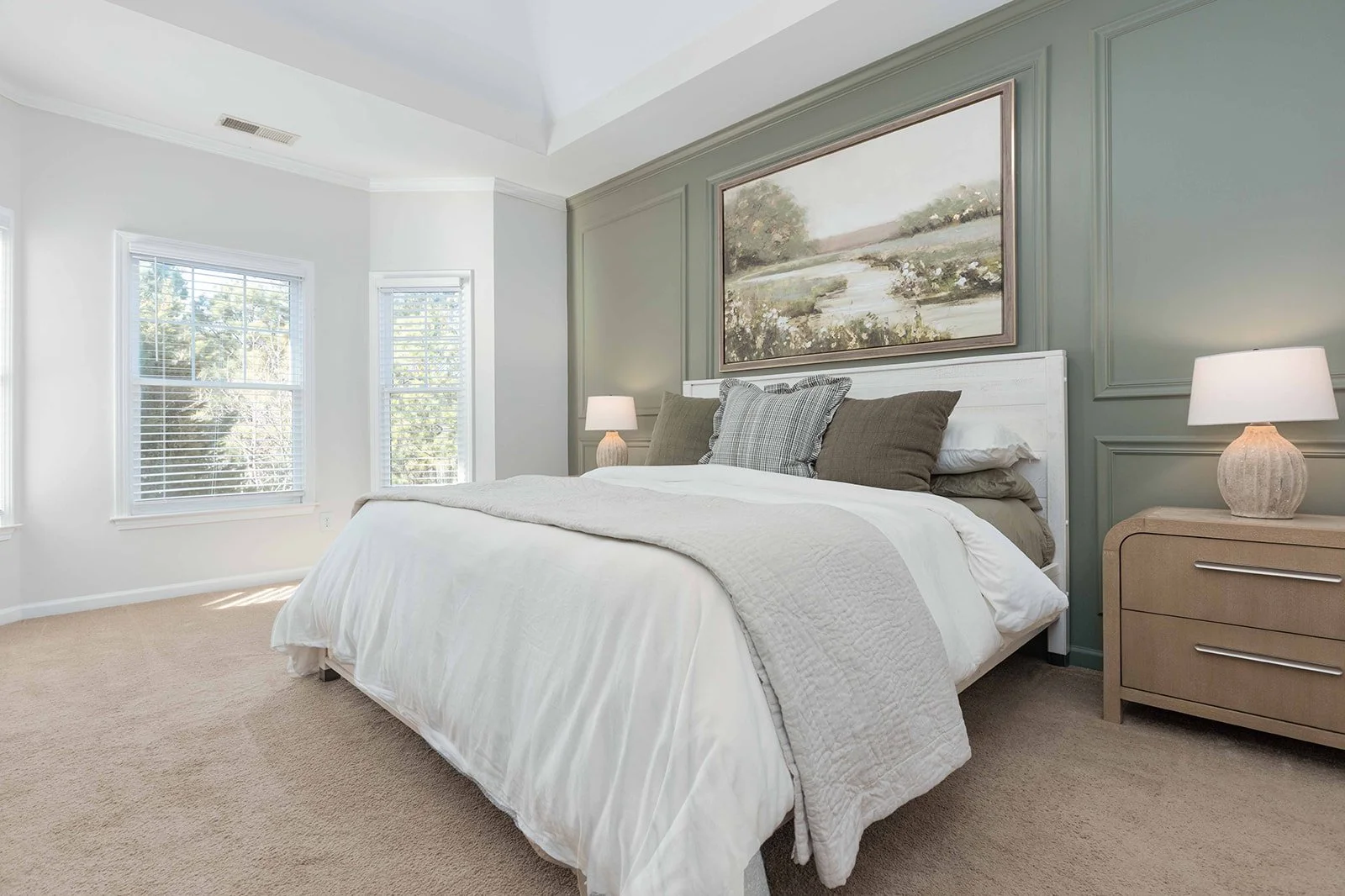 A bedroom with a white bed, beige nightstands with lamps, green and white walls, and large windows with blinds.
