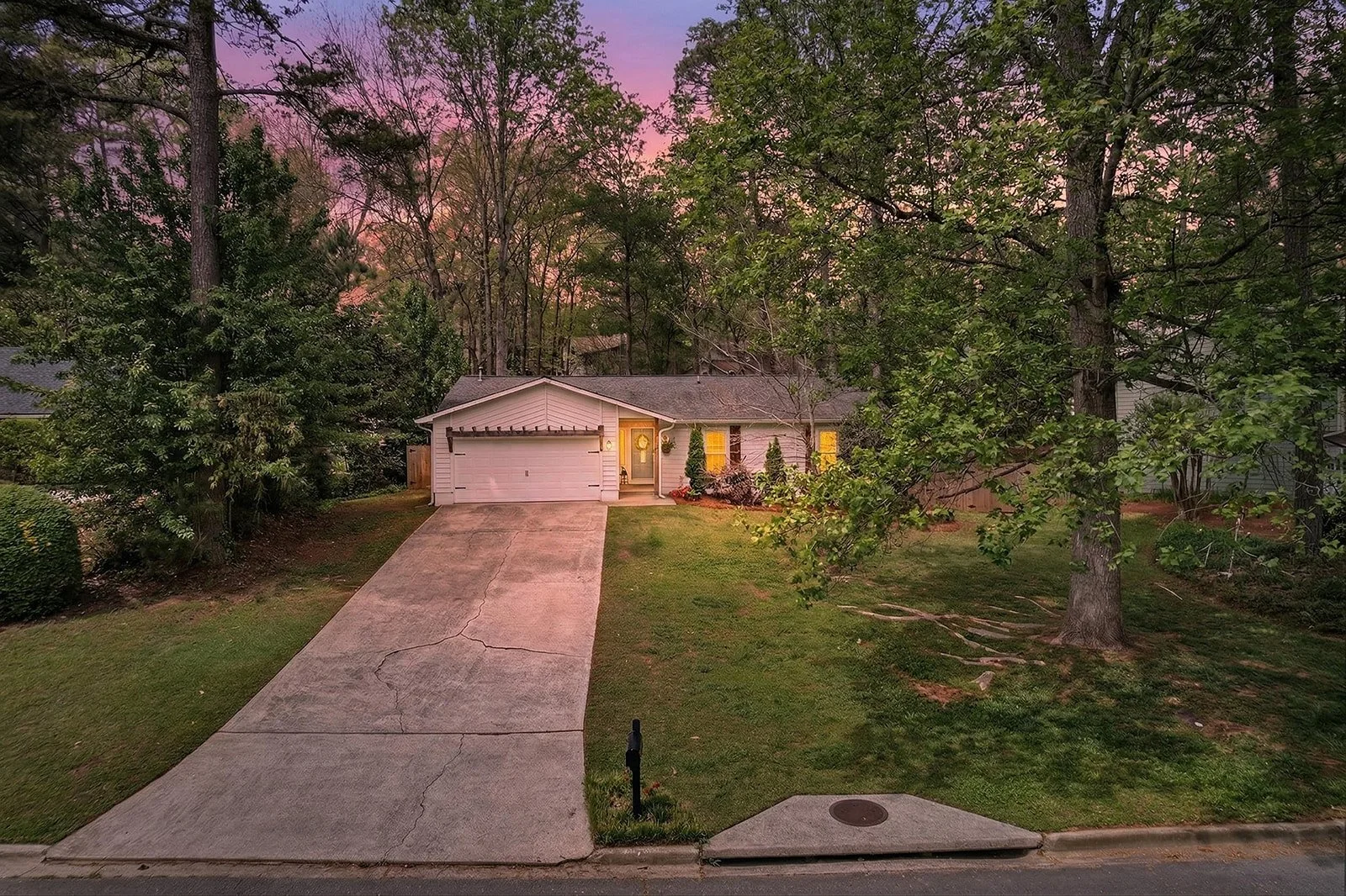 Front view of a house with a driveway, surrounded by trees and a lawn at sunset, with warm lighting glowing from windows.
