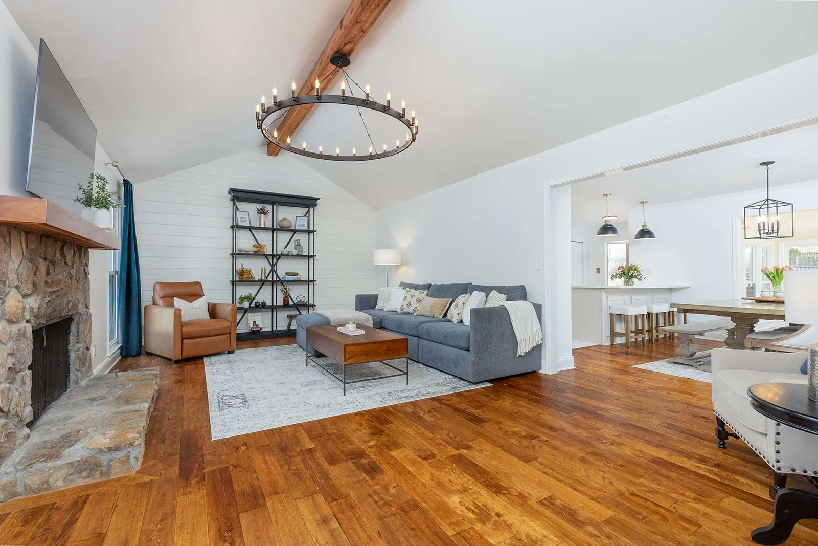 Living room with a gray sofa, wooden coffee table, leather armchair, stone fireplace, and a rug, connected to a dining area with a large wooden table, bench seating, and black pendant lights, all with hardwood floors and white walls.