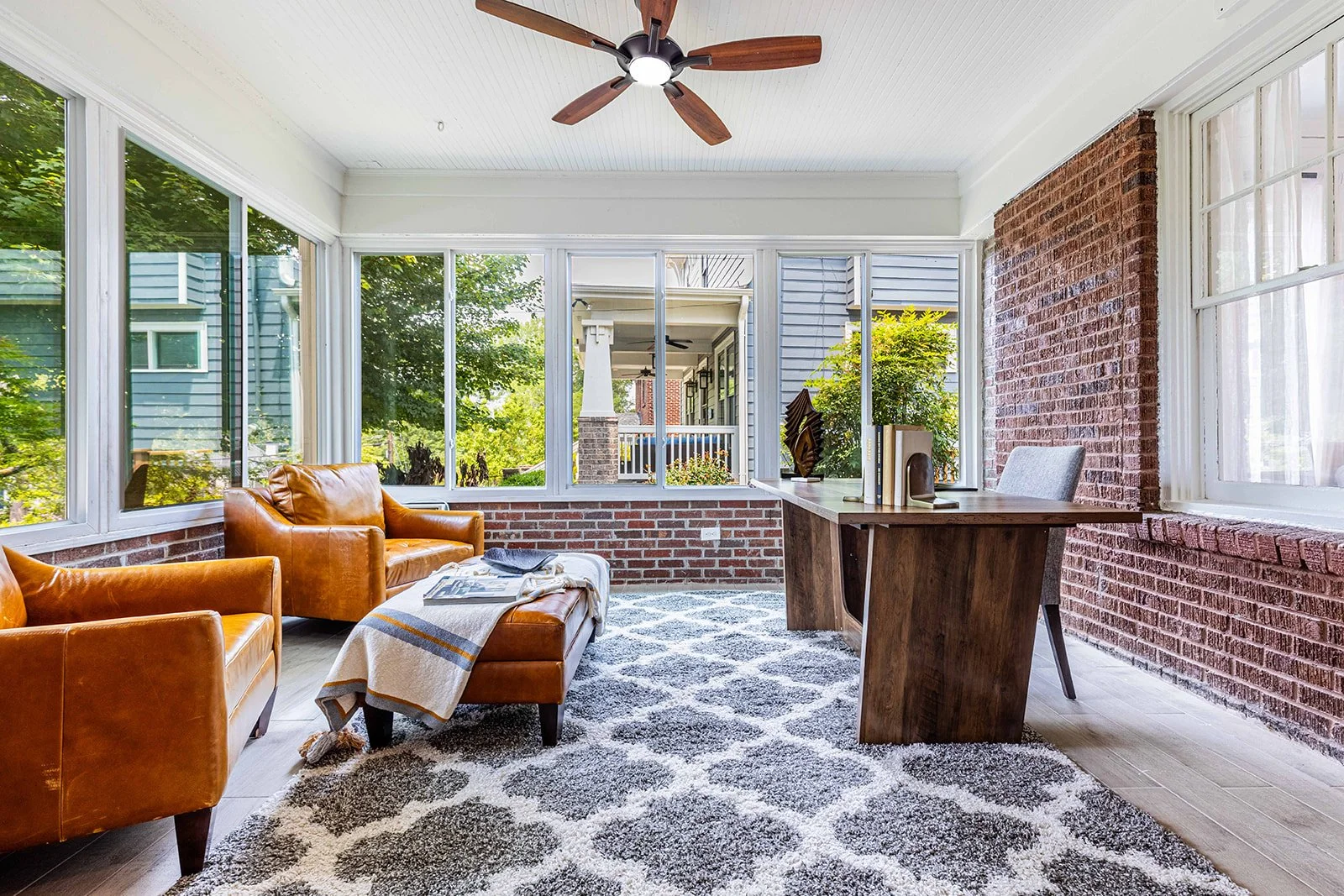 A sunroom with large windows, leather sofas, a wooden table, and a brick wall, decorated with a patterned rug and potted plants.