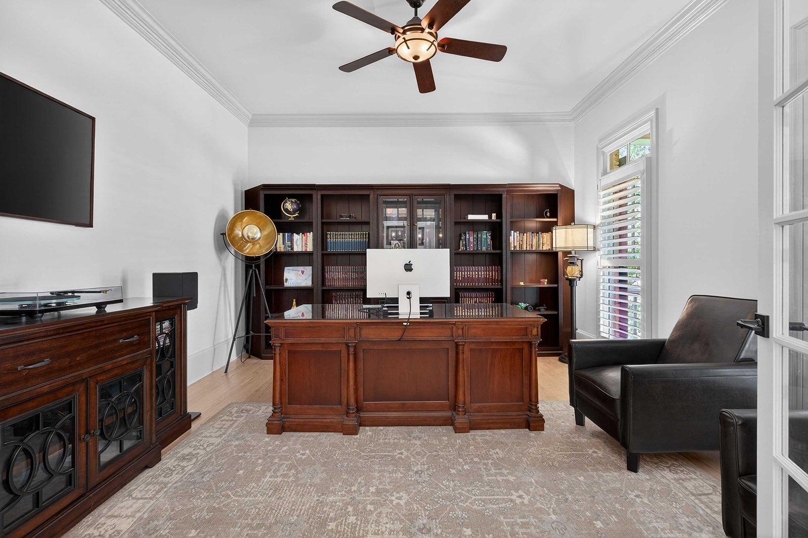 Home office with wooden desk, black leather armchair, large dark wooden bookshelf, vintage floor lamp, and mounted TV.