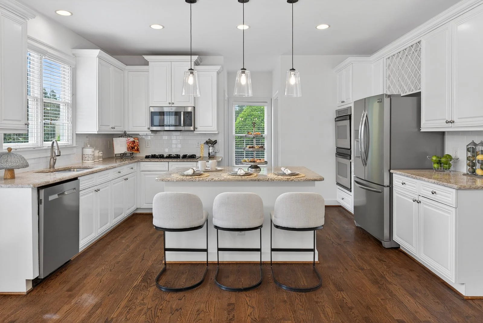 Modern kitchen with white cabinets, stainless steel appliances, a granite island with seating, hardwood floors, and natural light from windows.