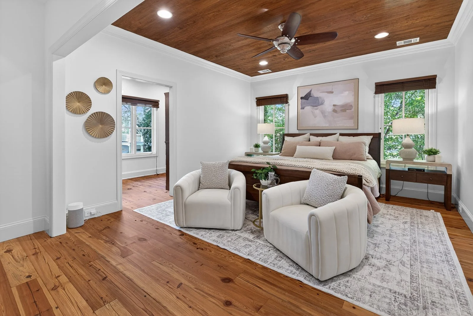 A bedroom with a wooden ceiling, white walls, and hardwood floors. The bed has a wooden headboard, white and beige bedding, and is flanked by two lamps on side tables. There are two white armchairs with pillows in front of the bed, and a decorative r