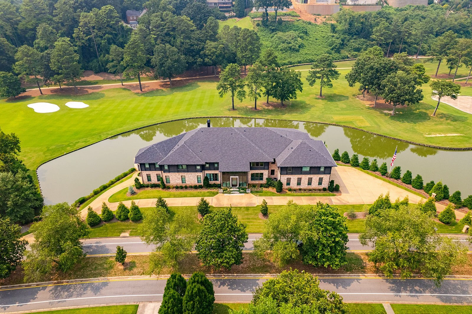 A large house situated near a small lake, surrounded by green trees and lawns, with a golf course nearby, and an American flag in the yard.