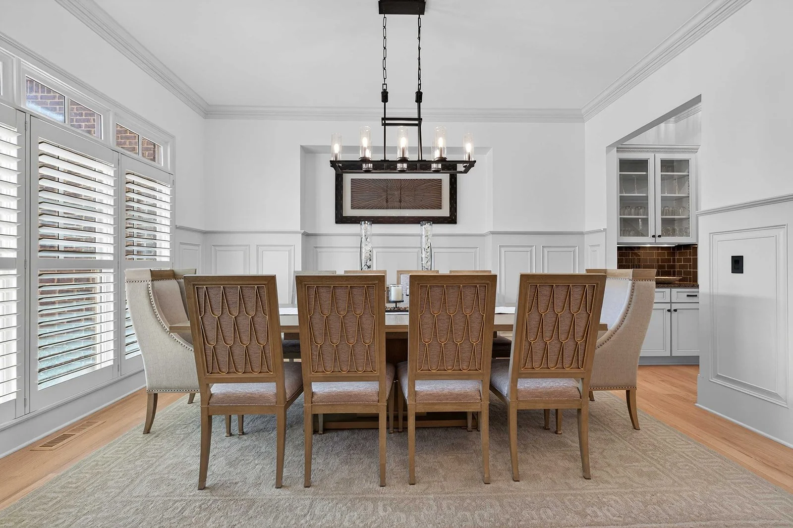 A dining room with a large rectangular table surrounded by ten chairs, with a chandelier above. The walls are white with crown molding. There are window shutters on the left, built-in cabinets on the right, and a framed artwork on the wall.