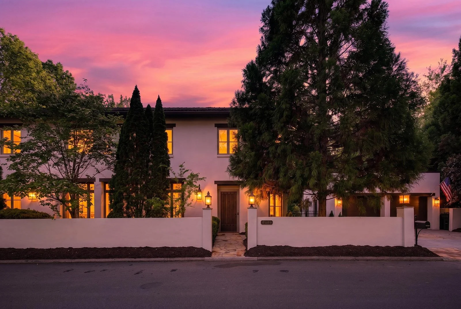 A modern two-story house during sunset with lit windows, white exterior walls, a wooden front door, and a front yard fenced by a white wall. Tall trees and bushes surround the house, and the sky is pink and purple.
