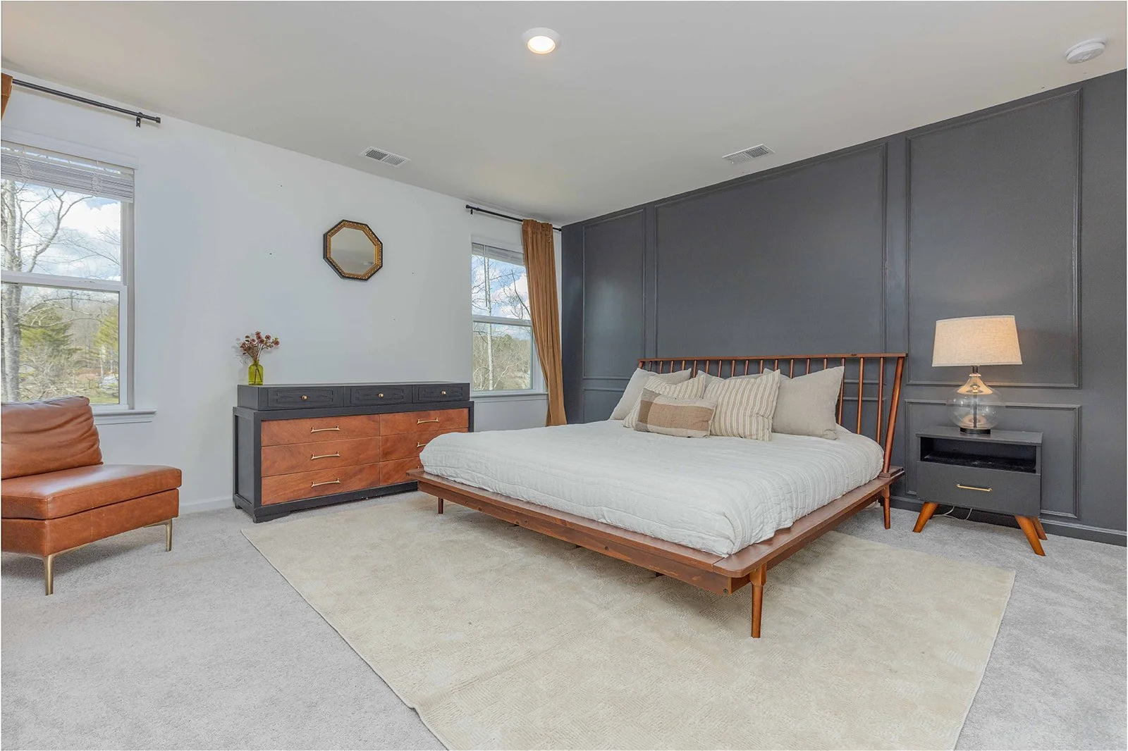 Modern bedroom with a large bed on a wooden frame, a dark gray nightstand with a table lamp, a dresser with a flower vase, a chest of drawers, a leather chair, and large windows with curtains providing natural light.