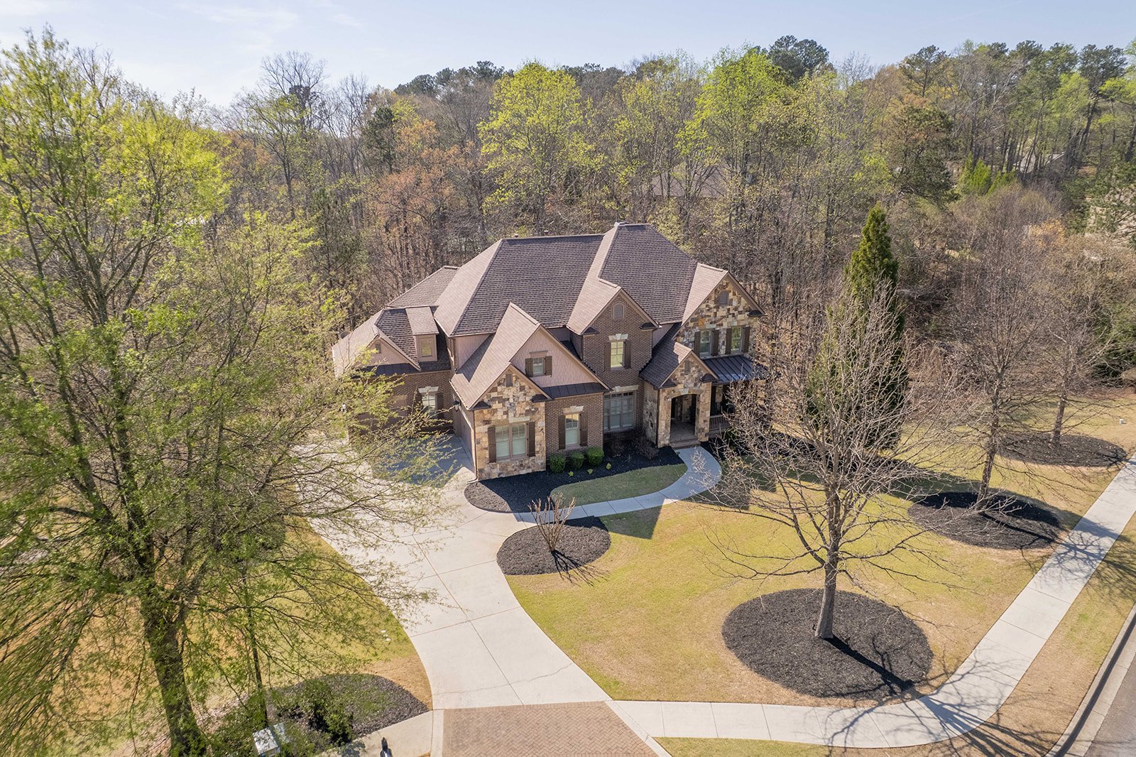 Aerial view of a large brick house with a multi-gabled roof, surrounded by a landscaped yard with trees, pathways, and a lawn, in a suburban neighborhood.