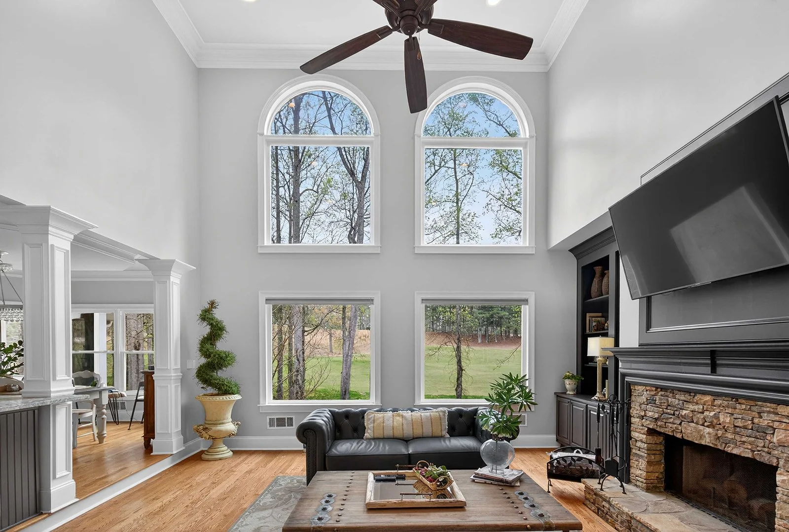 Living room with high vaulted ceiling, three large arched windows showing trees outside, a ceiling fan, a black leather sofa with a striped pillow, a fire place with brick surround, a flat-screen TV mounted above, a black built-in shelf with decorati