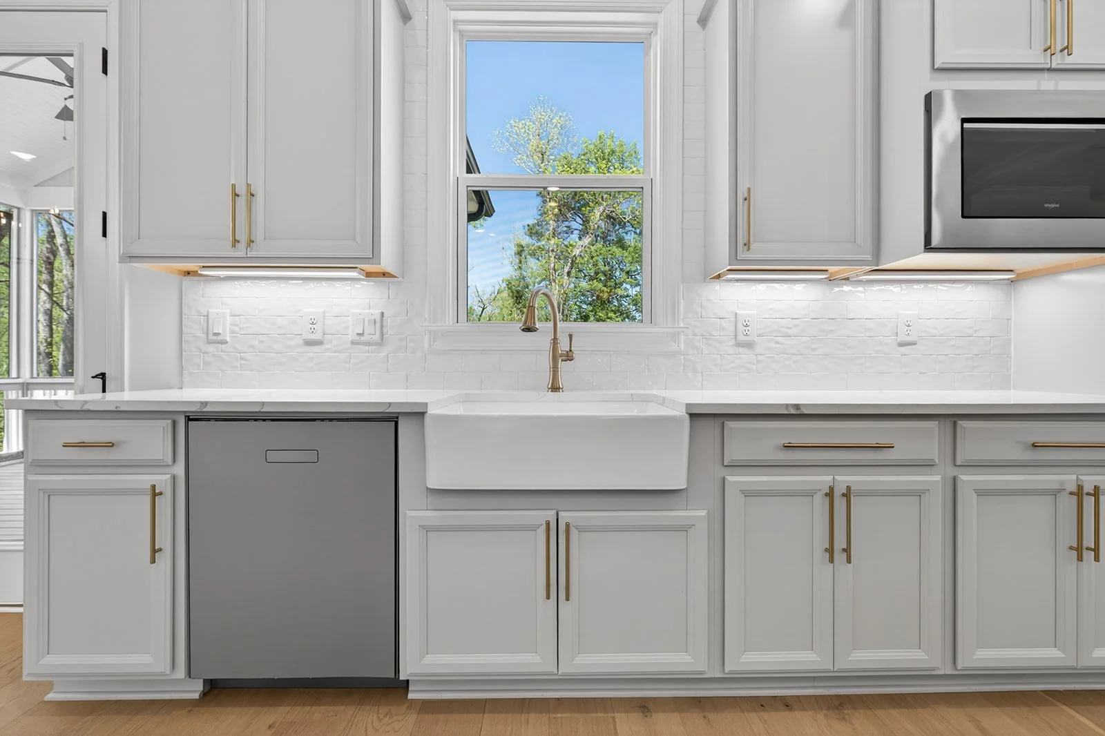 Modern white kitchen with a farmhouse sink, gold handles on cabinets, and a window showing trees outside.